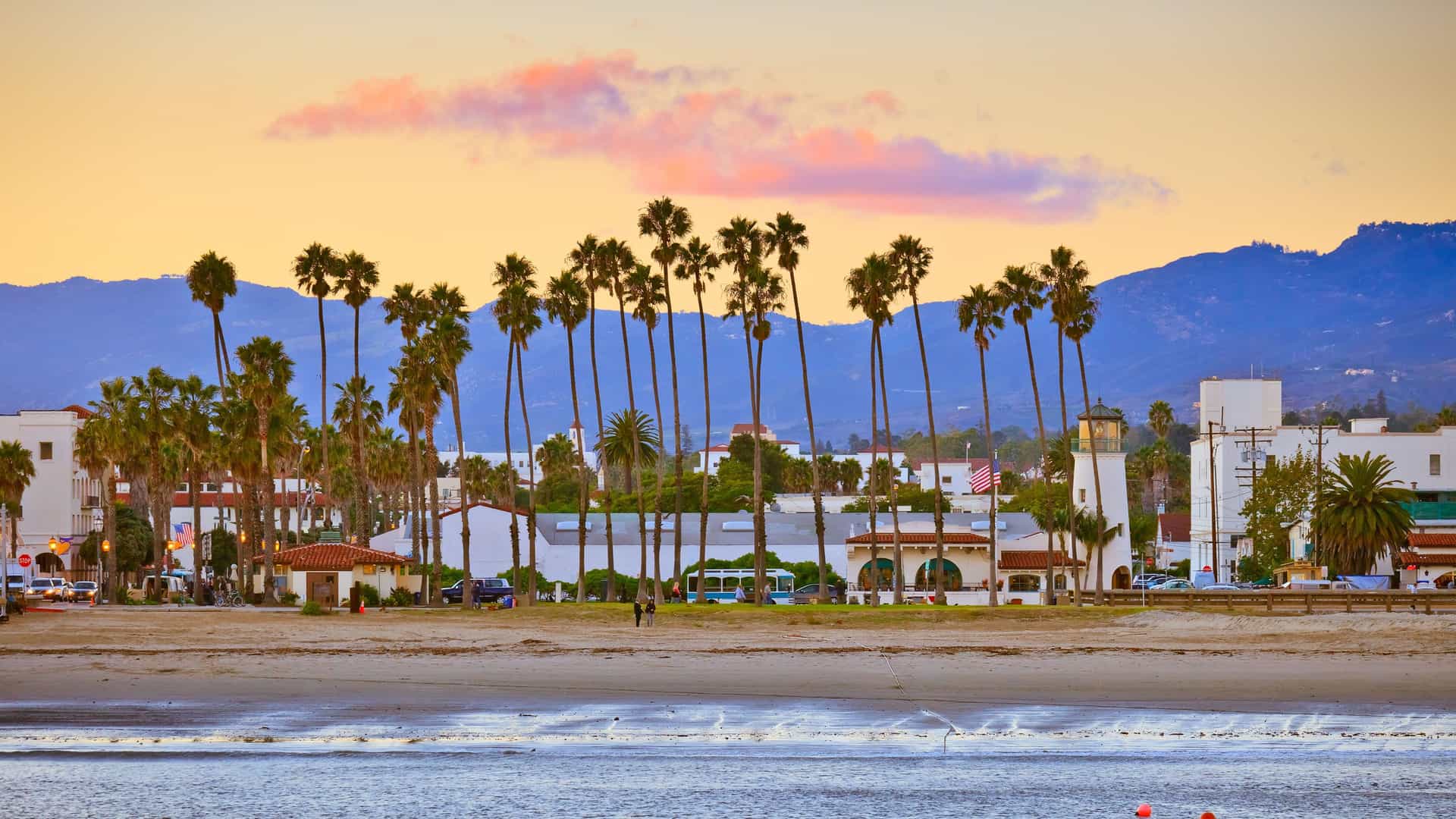 A serene sunset view of a beautiful sandy beach in Santa Barbara, California, with a row of iconic palm trees and the coastal cityscape with mountains in the background.