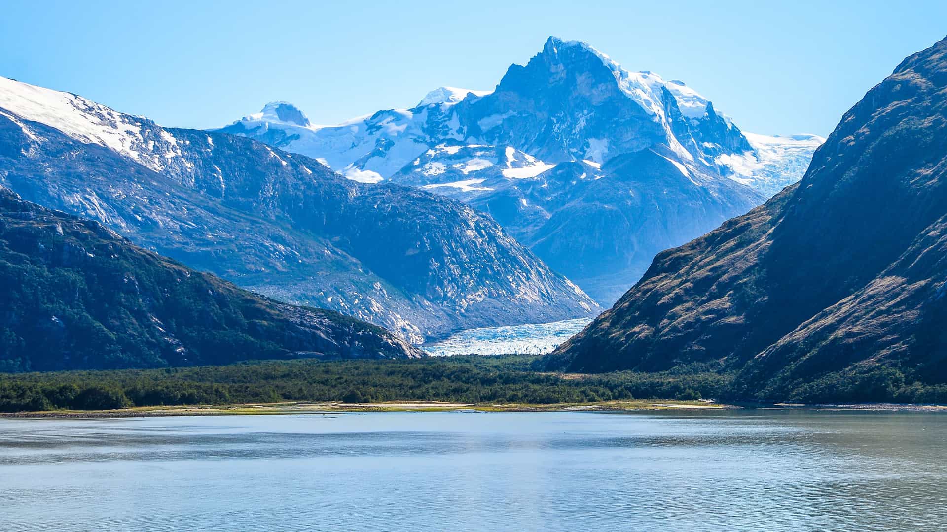 A breathtaking landscape of Seno Eyre Fjord in Chilean Patagonia, with a glacier nestled between towering, snow-capped mountains and a calm body of water in the foreground.
