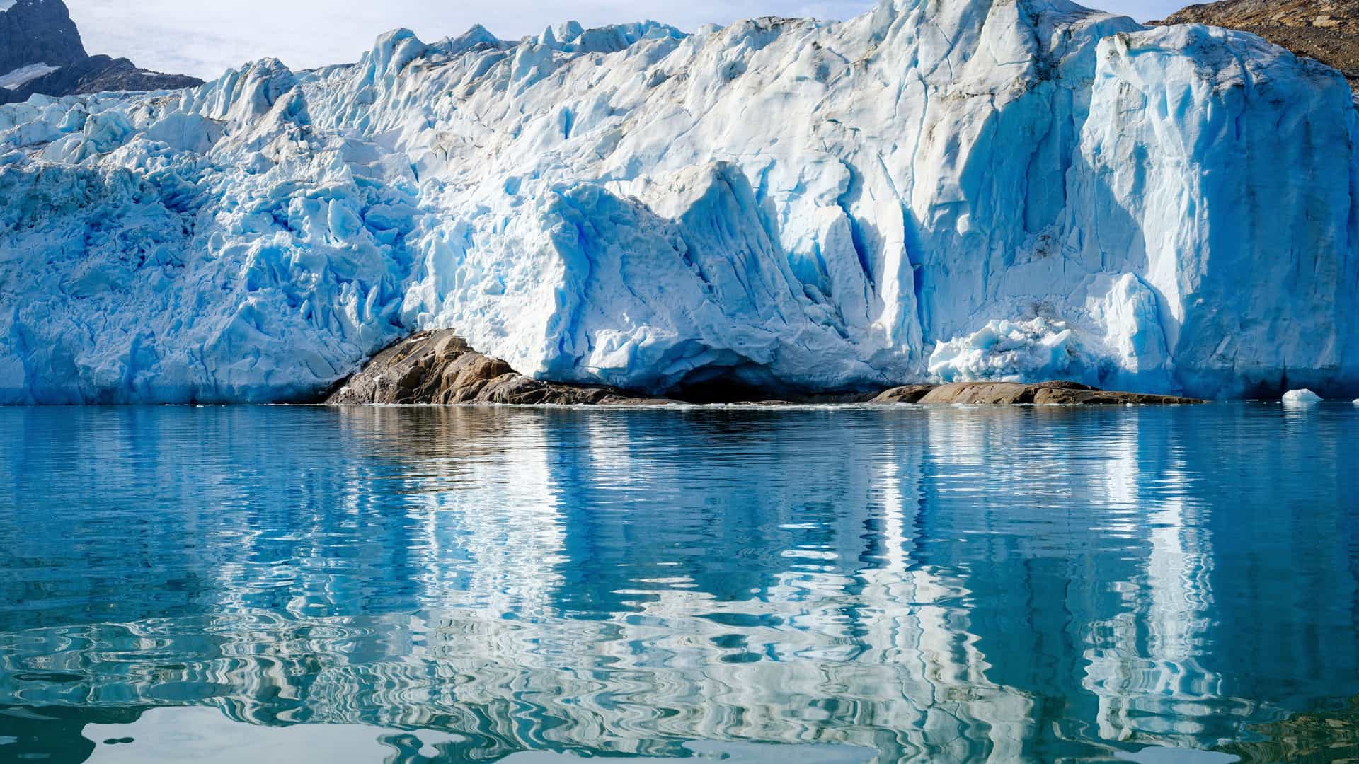 A majestic view of a massive, blue Sermiligaaq glacier in Greenland, with its large ice formations and deep crevasses reflected in the calm, icy water of the fjord.