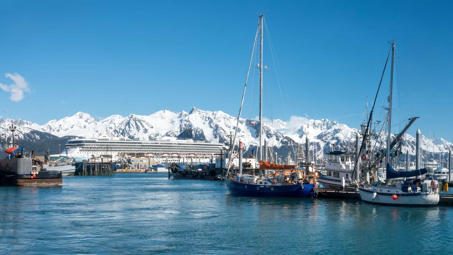 "A large cruise ship and several sailboats docked in the harbor of Seward, Alaska, with the Chugach Mountains' snow-covered peaks visible in the distance.  "