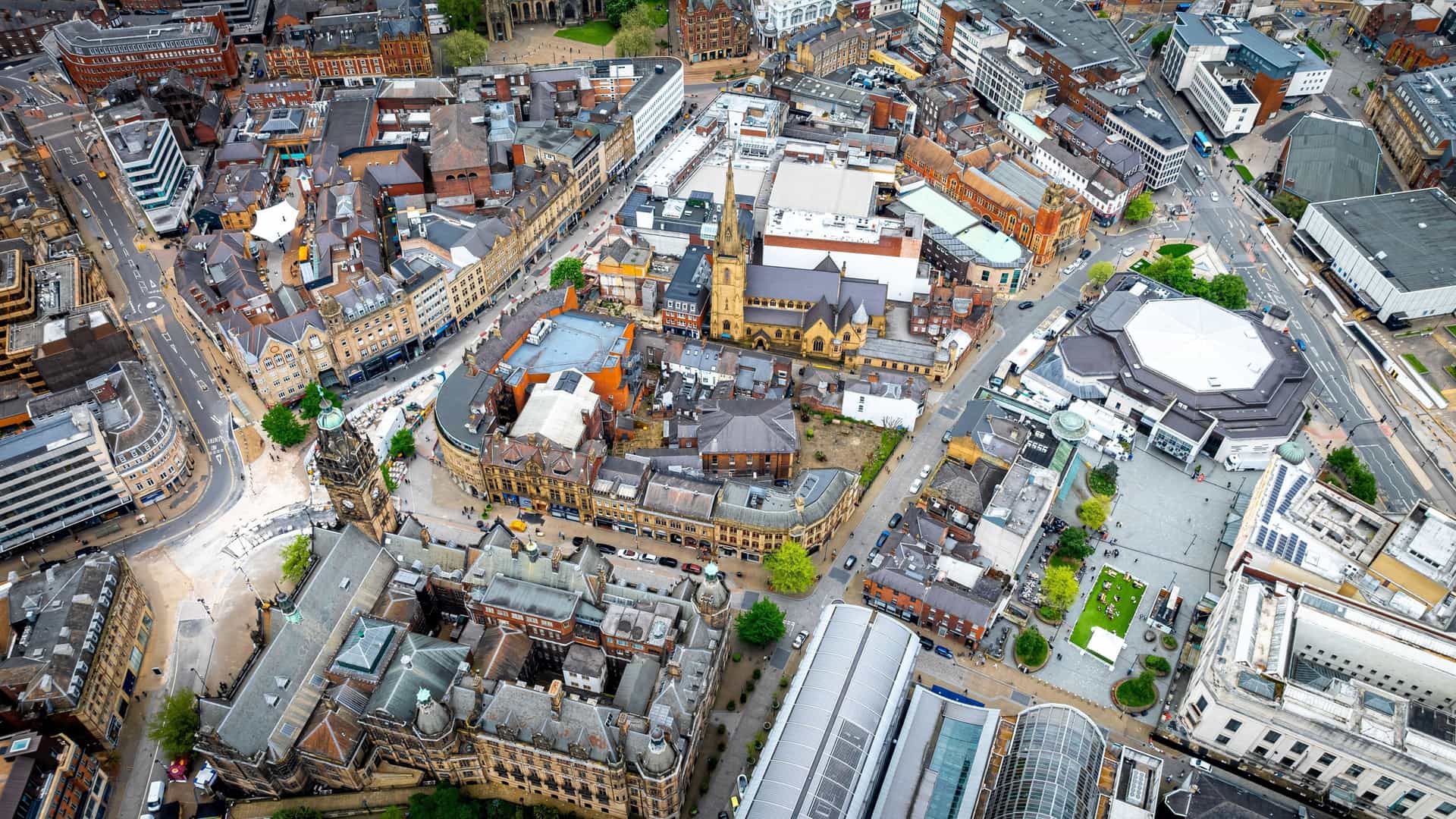 An aerial, panoramic view of the Sheffield cityscape in the English county of South Yorkshire, UK. The image captures the dense urban center with its modern high-rise buildings and historic architecture (potentially including landmarks like the Arts Tower or Town Hall), set against the city's famously green, hilly surroundings which often extend towards the Peak District National Park.