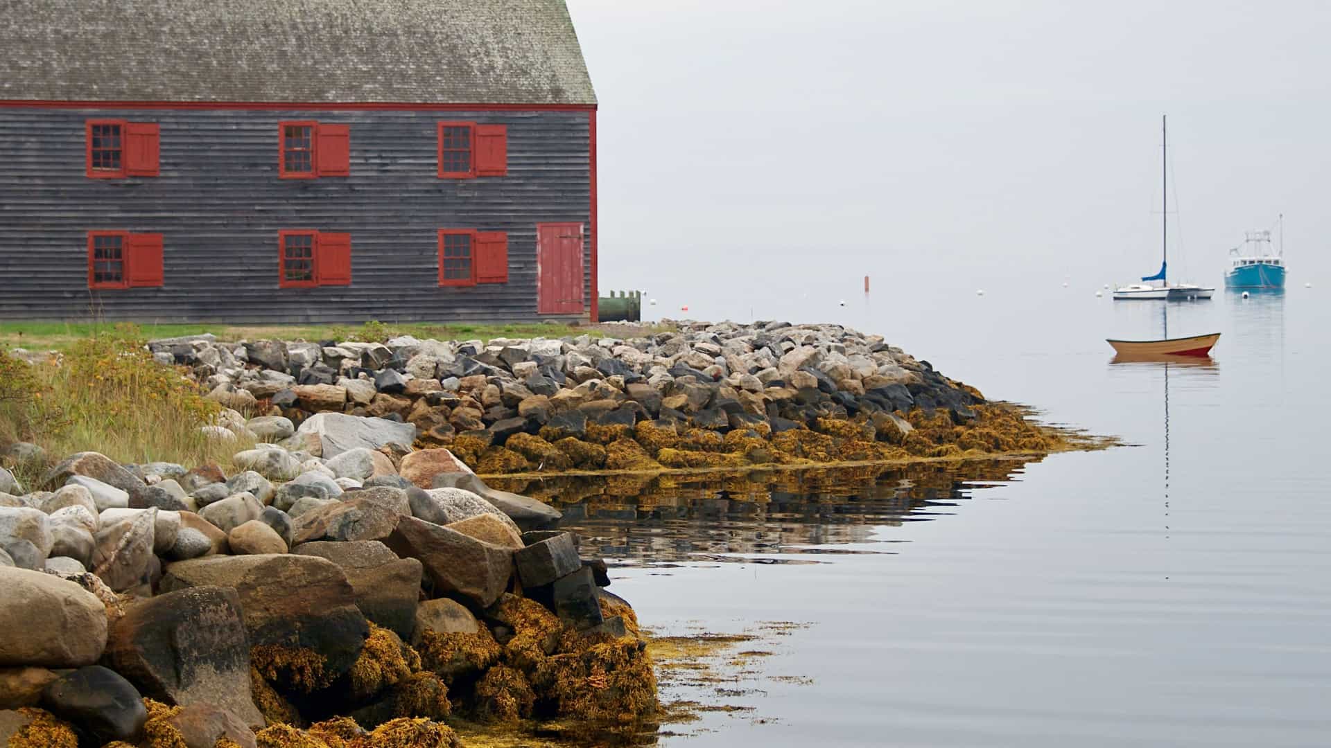 A scenic view of a rustic, grey clapboard building with red shutters on the rocky shoreline of Shelburne, Nova Scotia, with small boats anchored in the calm water.
