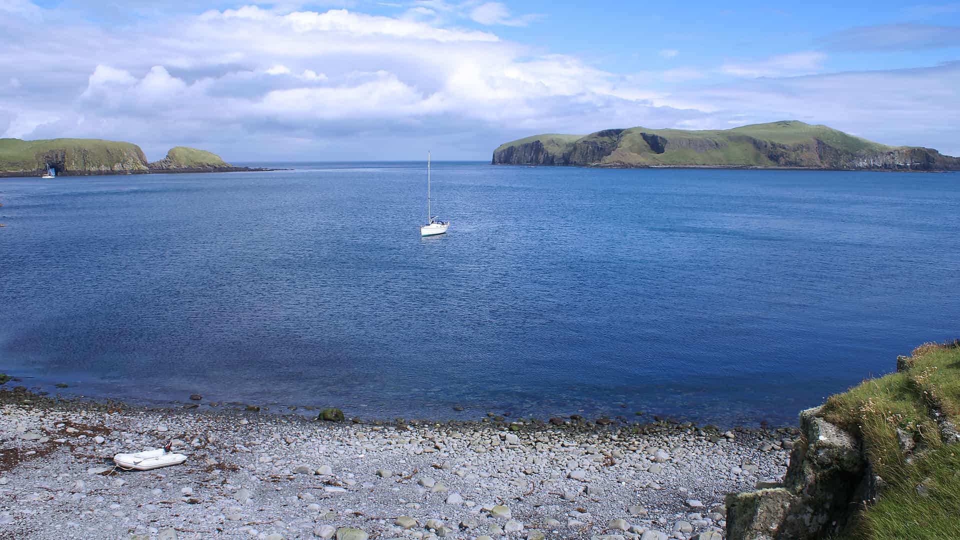 A beautiful wide-angle view of the scenic coastline of the Shiant Islands, Scotland, with a sailboat anchored in the bay and a pebble beach in the foreground.