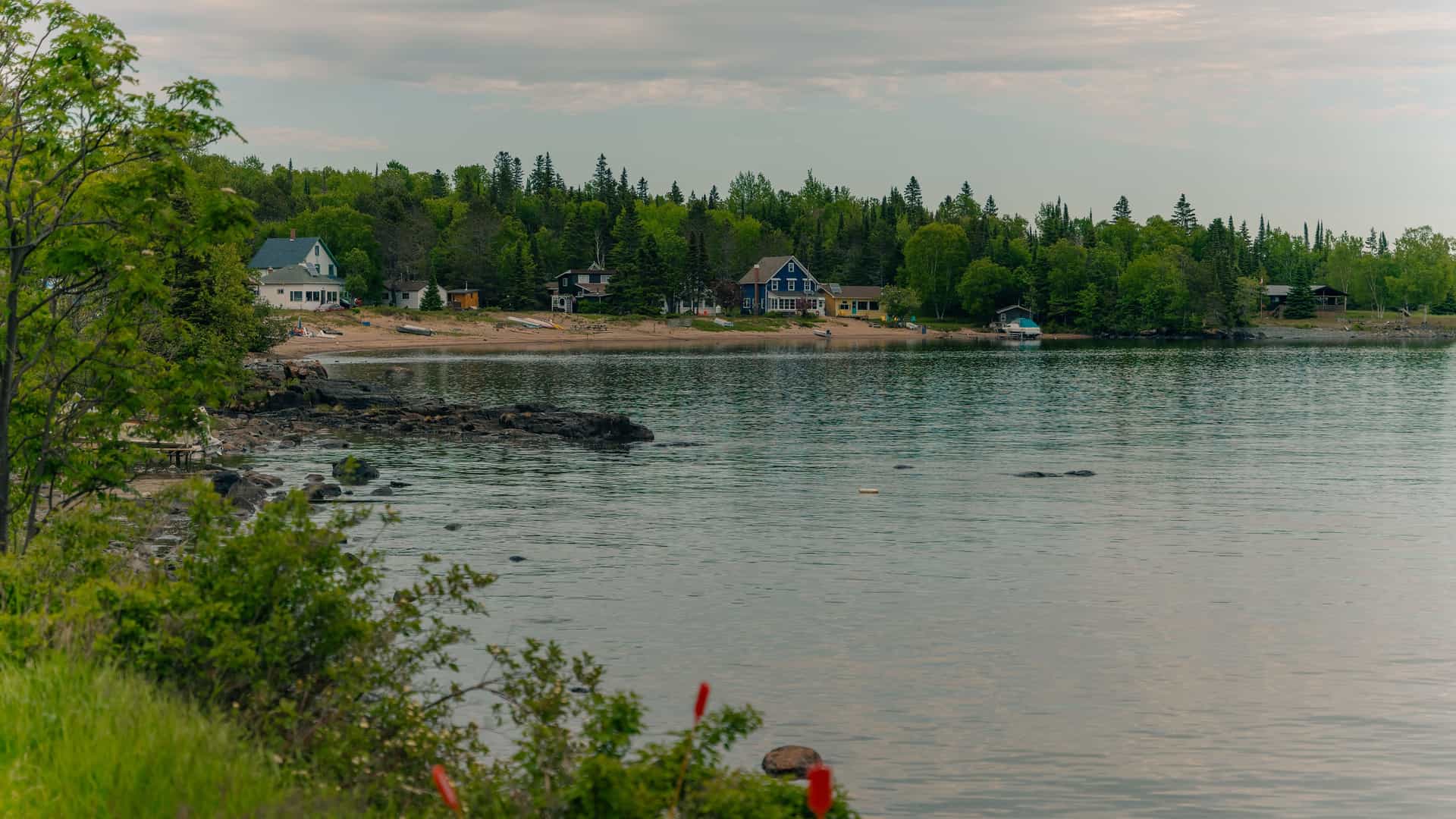 A tranquil view of Silver Islet, Canada, with a few colorful cottages and docks along the rocky shoreline of Lake Superior, surrounded by lush green trees and a quiet sky.
