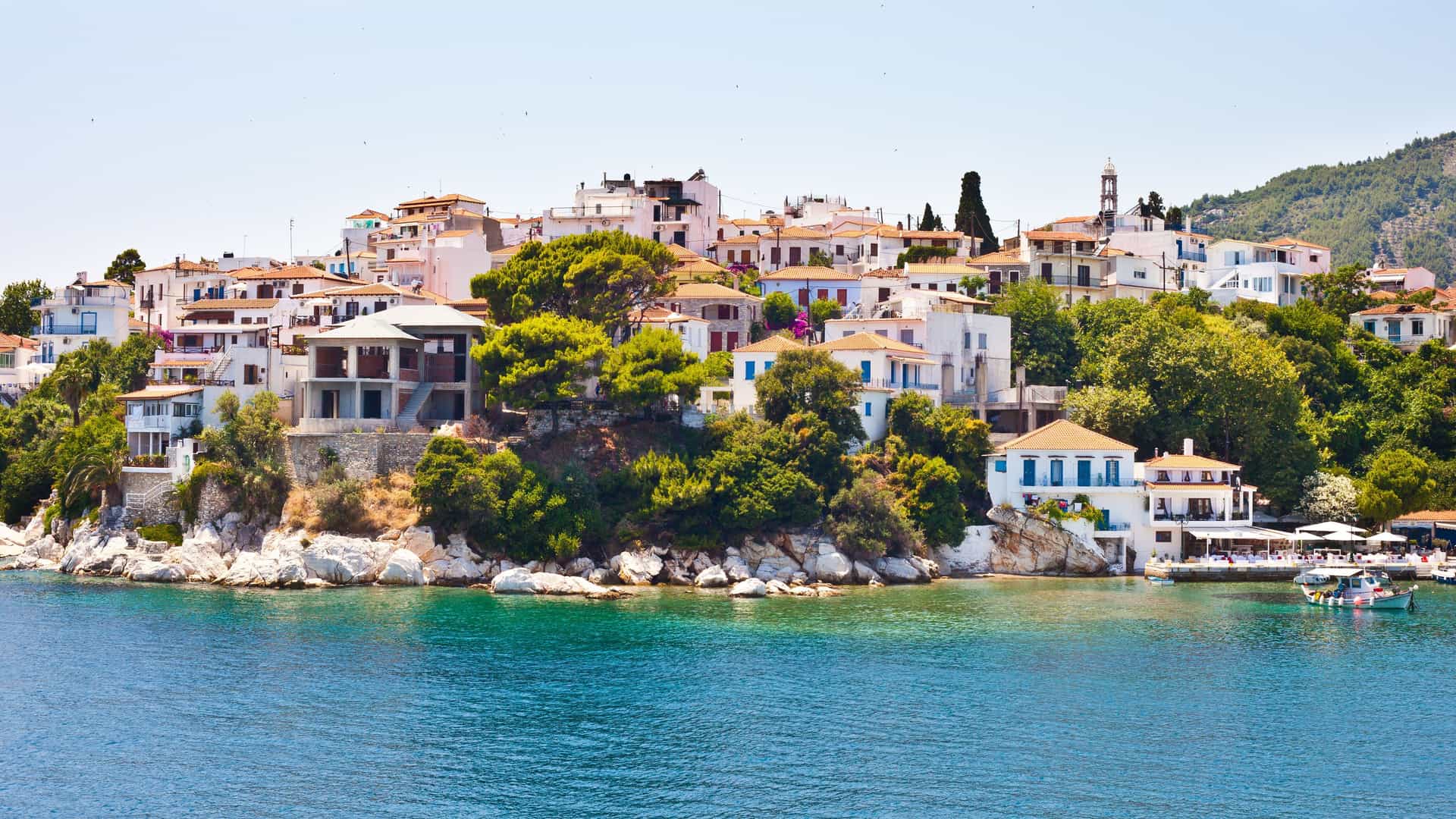 A stunning daytime shot of Skiathos Town, with its traditional white-washed houses and red-tiled roofs nestled on a hillside above the sparkling blue Aegean Sea.