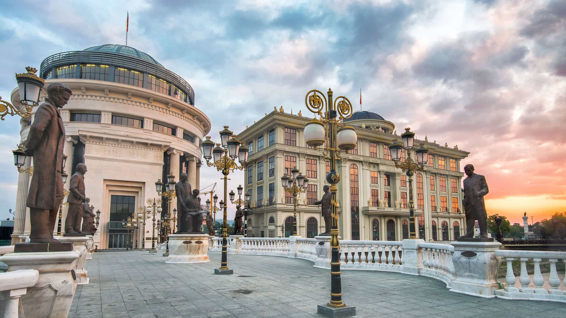 A beautiful sunset view of the Vardar River in Skopje, Macedonia, with the impressive Museum of the Macedonian Struggle and Archaeological Museum buildings and numerous statues on the bridge.