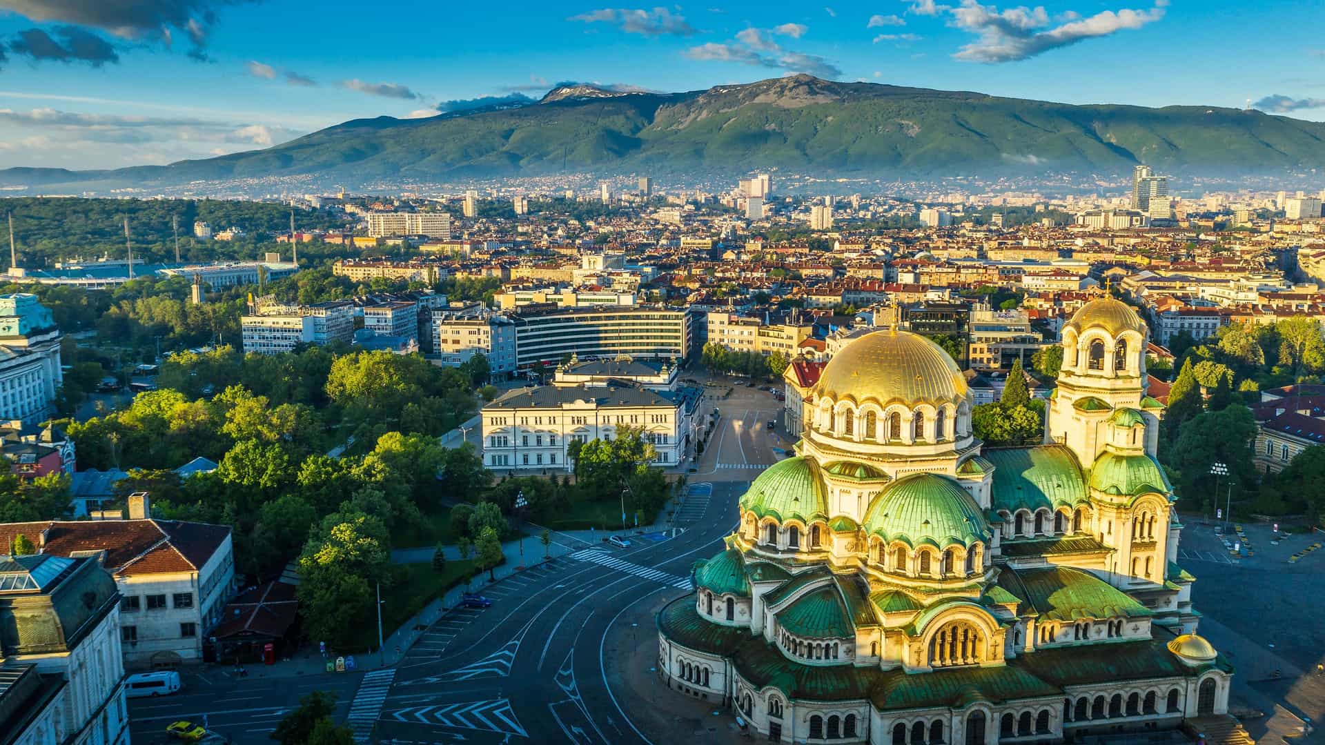 A stunning high-angle view of the iconic Alexander Nevsky Cathedral in Sofia, Bulgaria, with its green and gold domes, set against the backdrop of the city and Vitosha Mountain.