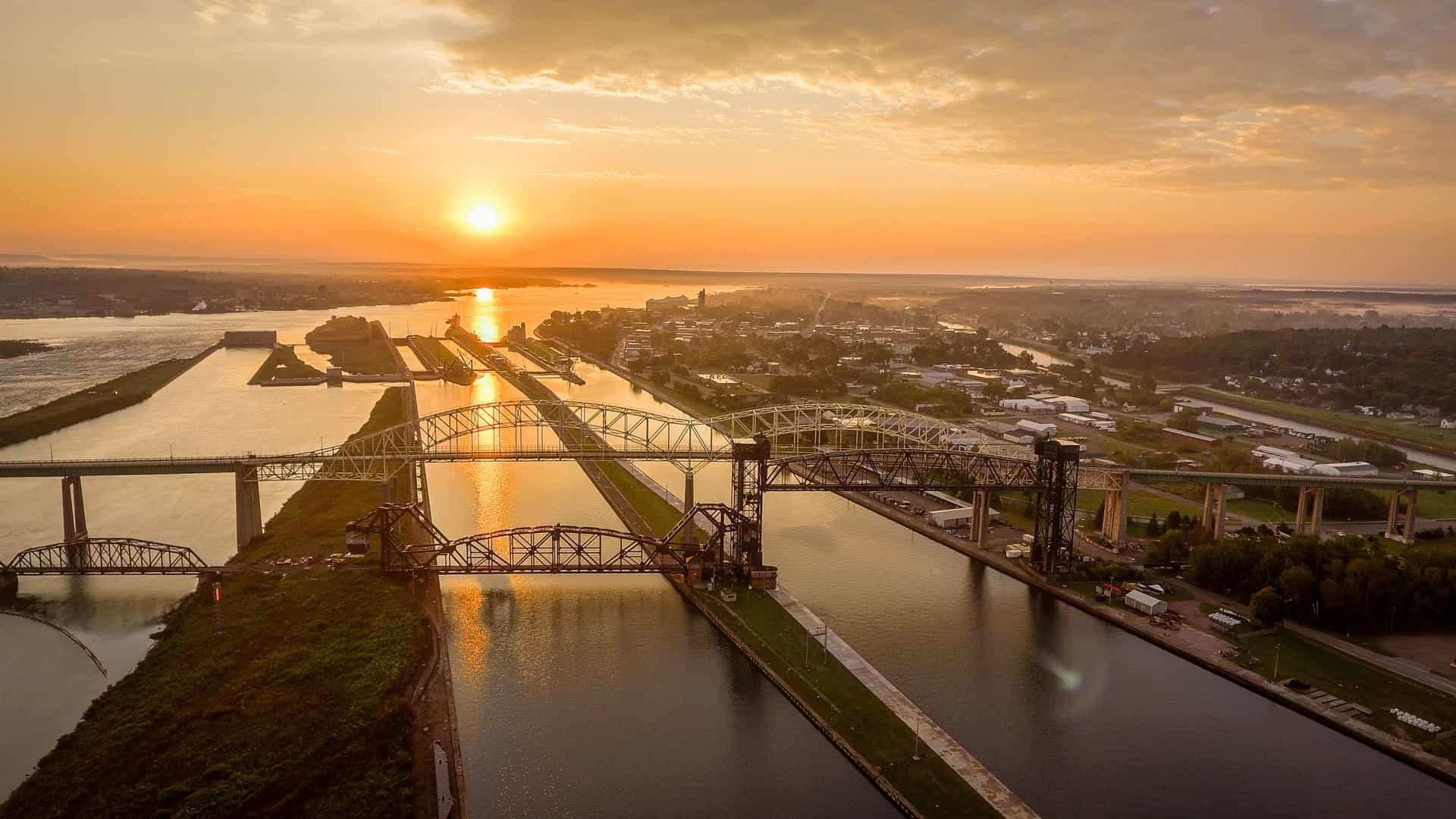 An aerial view of the Soo Locks in Sault Ste. Marie, Michigan, with multiple bridges crossing the canals, and the sun setting over the tranquil water.