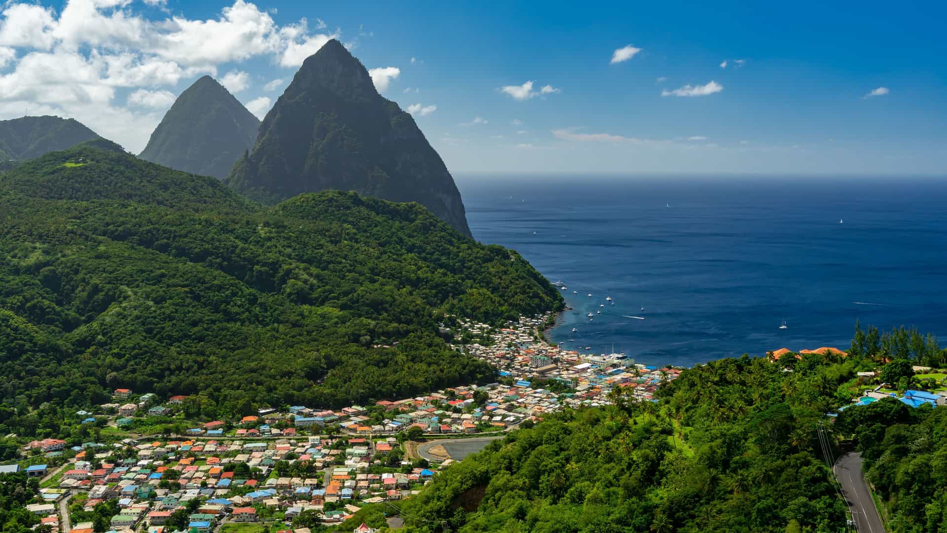 "A scenic view of the coastal town of Soufrière, St. Lucia, with its colorful houses nestled beneath the famous twin volcanic peaks, the Pitons, and the Caribbean Sea.  "