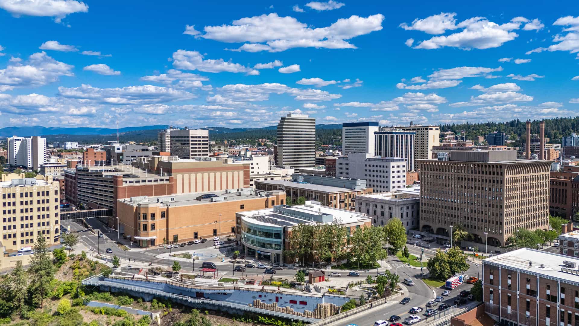 "An aerial view of the Spokane, Washington skyline, with its downtown buildings and the surrounding hilly landscape under a bright, cloud-filled blue sky.  "