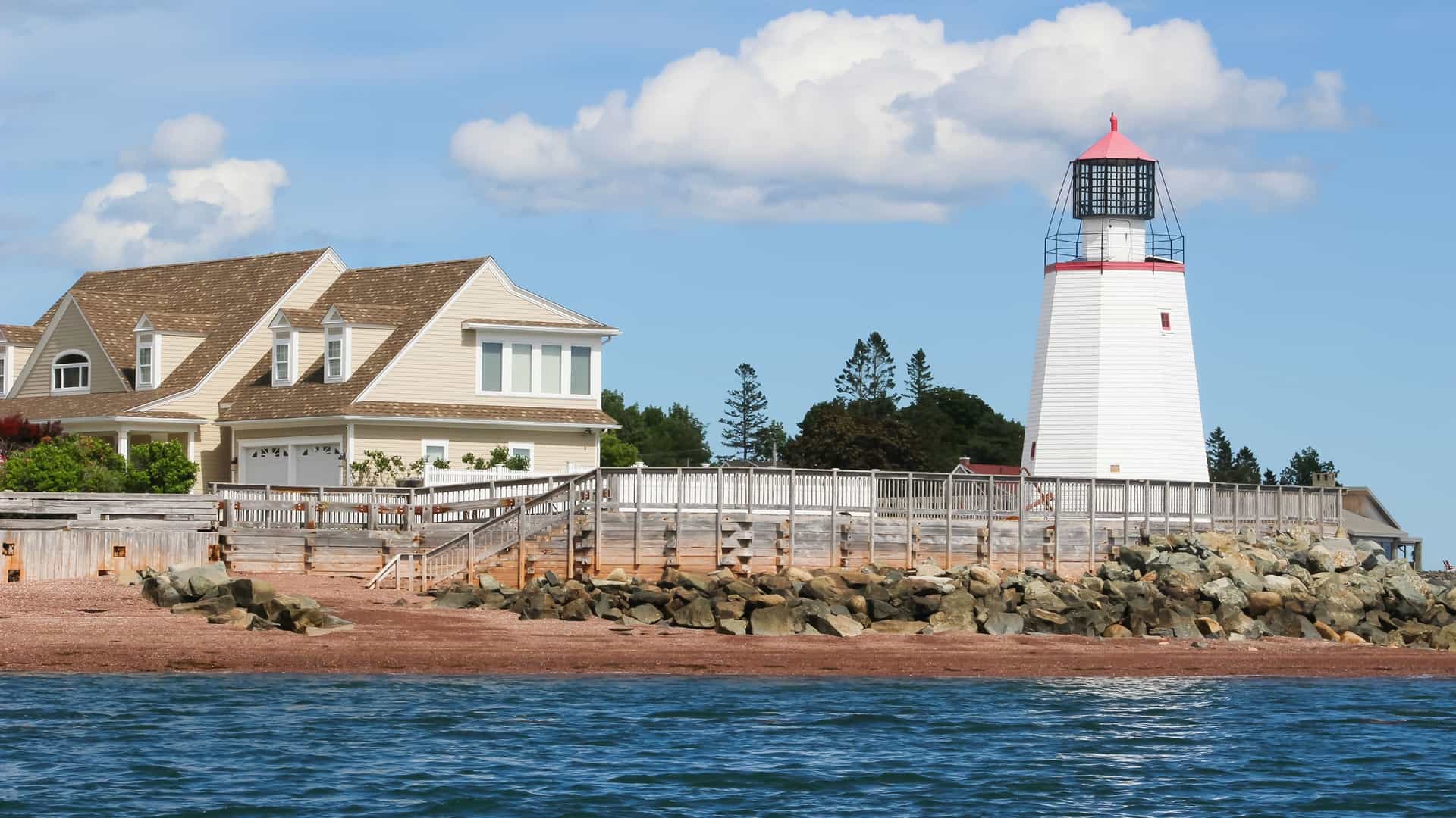 A white lighthouse with a red top stands next to a large, tan shingled house on a rocky, red-sand beach.