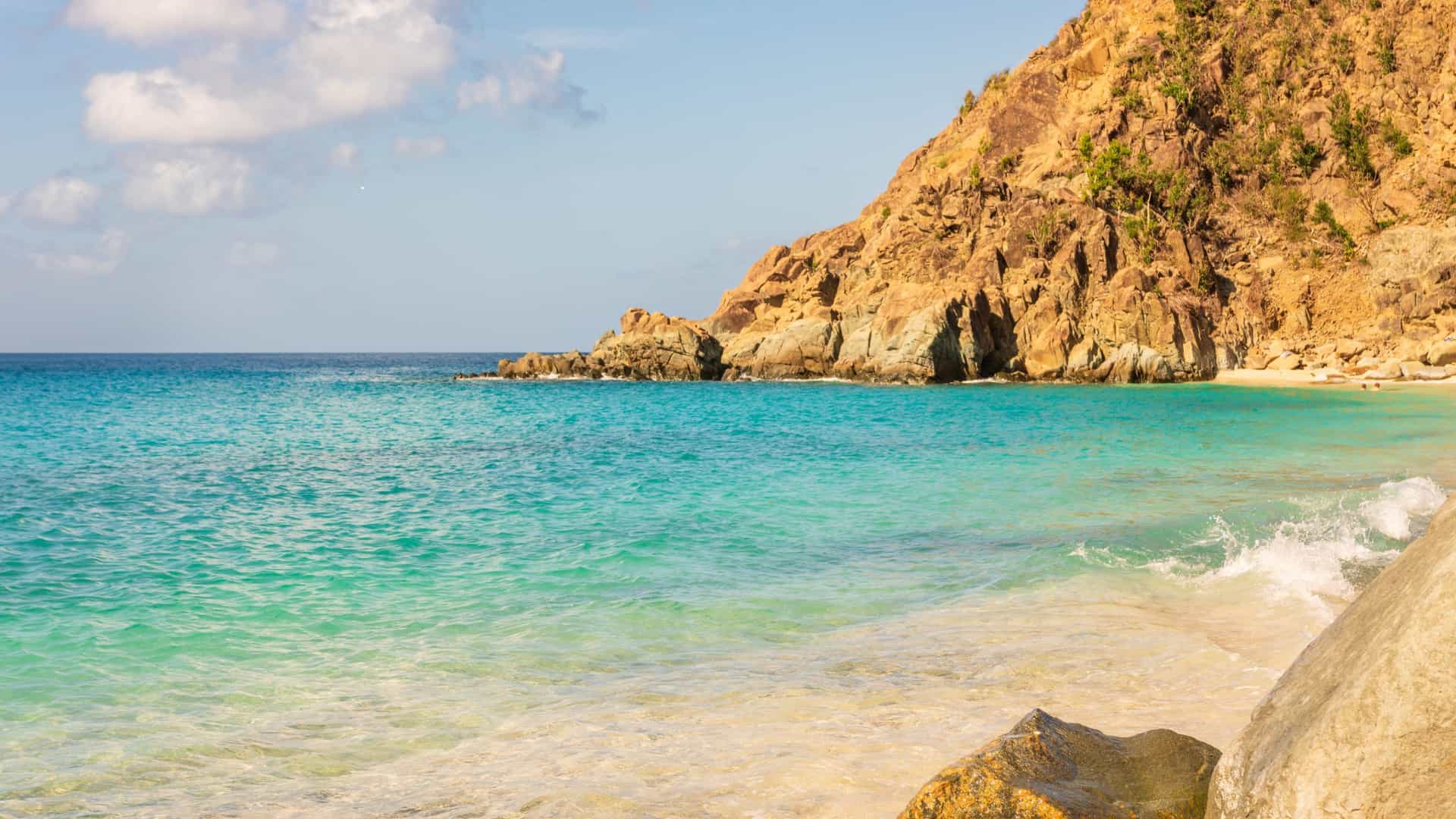 Crystal-clear turquoise water laps against a sandy beach at the foot of a tall, rugged orange cliff under a bright, sunny sky.