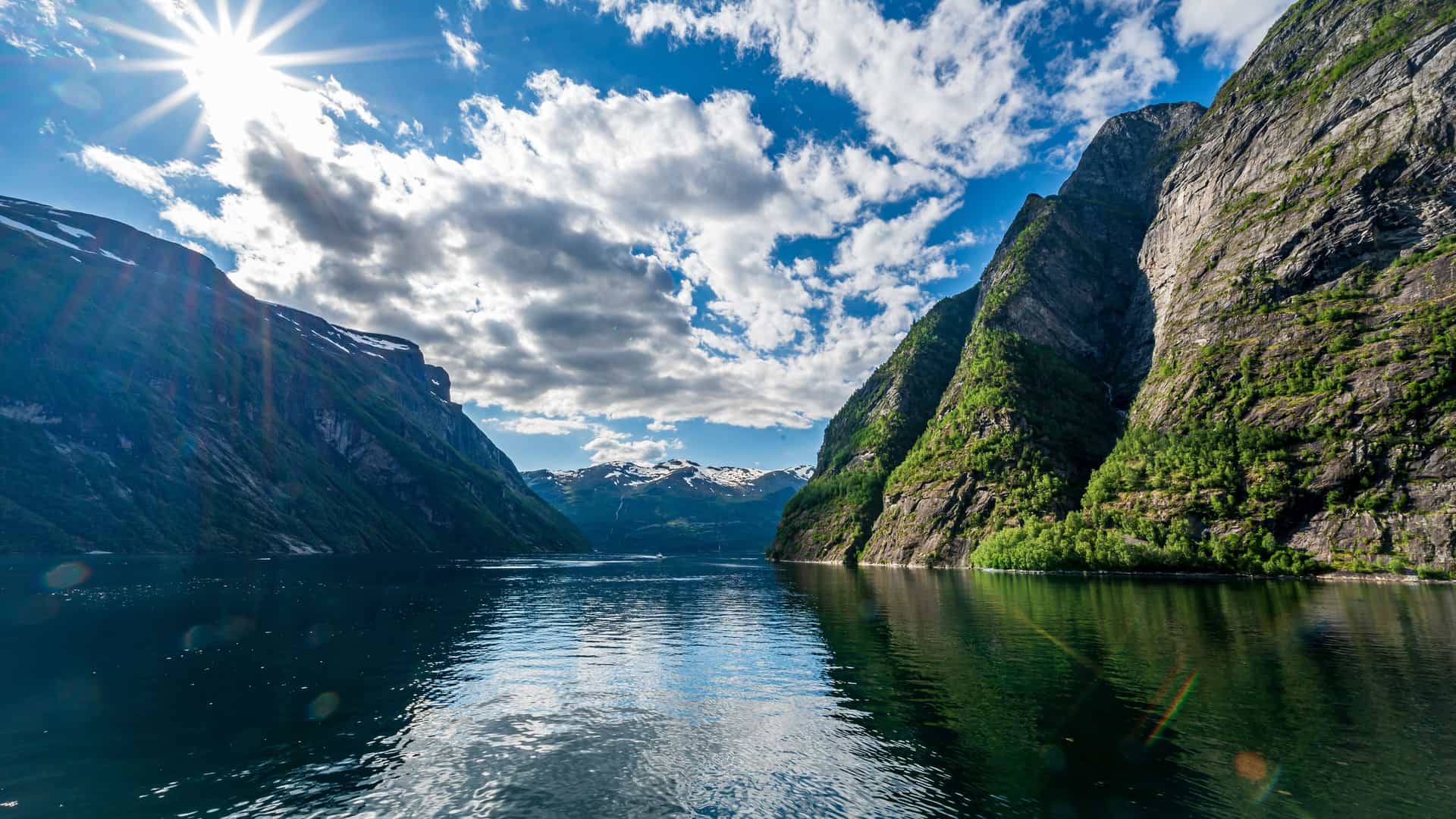 A scenic view of the majestic Storfjorden in Norway, with towering green mountains on both sides of a calm, reflective blue fjord under a bright, sunny sky.