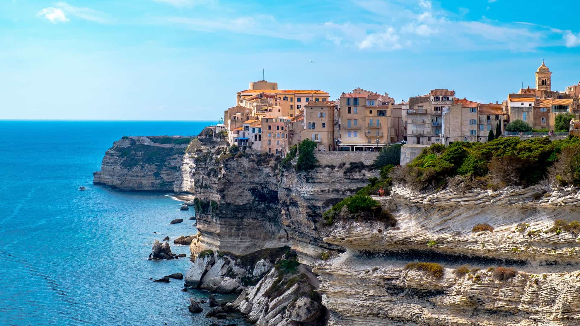 A panoramic view of the clifftop town of Bonifacio on the island of Corsica, with historic buildings perched on the dramatic white limestone cliffs overlooking the beautiful blue Strait of Bonifacio.