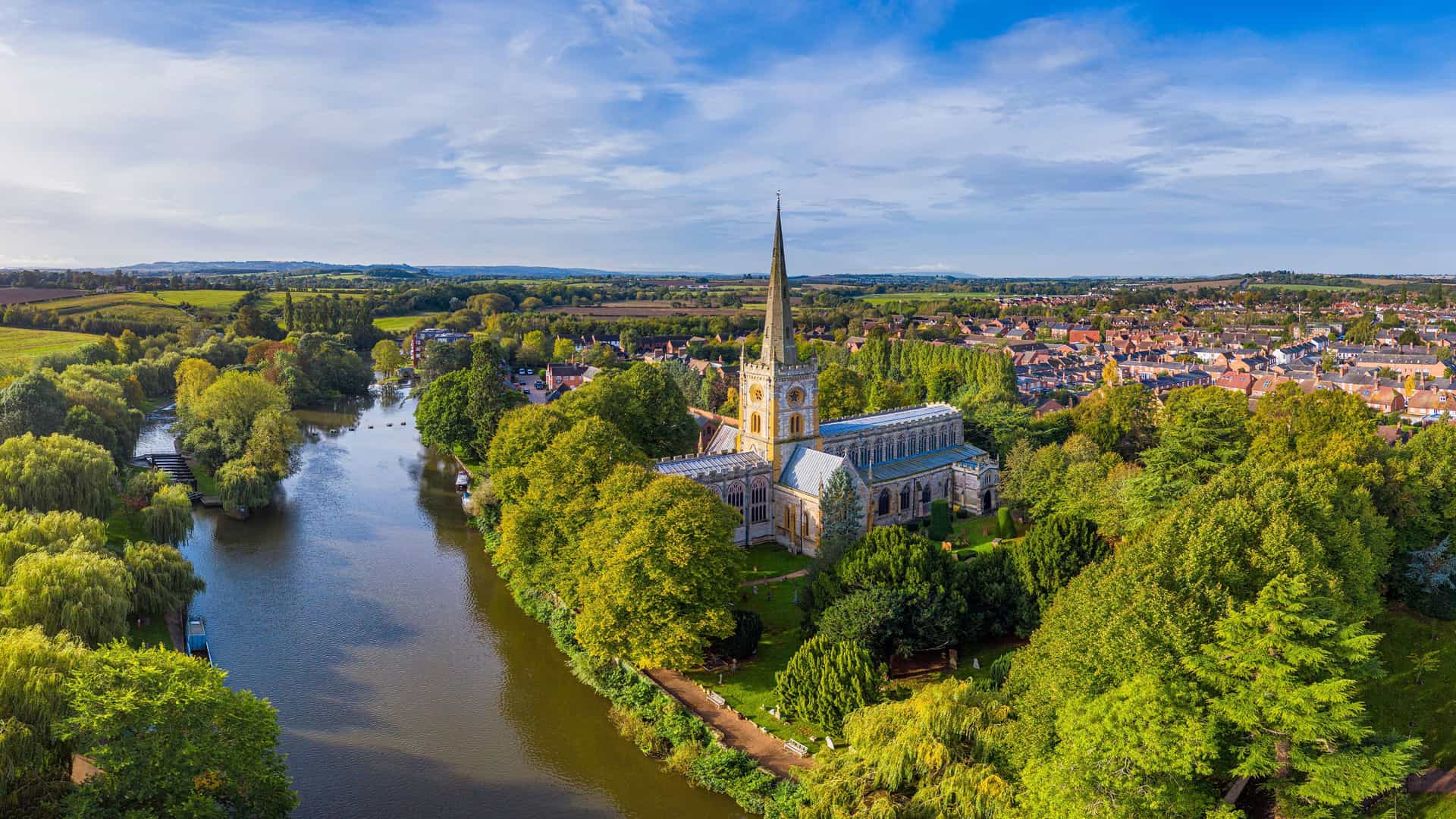 An aerial view of the historic Holy Trinity Church in Stratford-upon-Avon, England, nestled on the banks of the tranquil River Avon, surrounded by lush greenery and the town's rooftops.