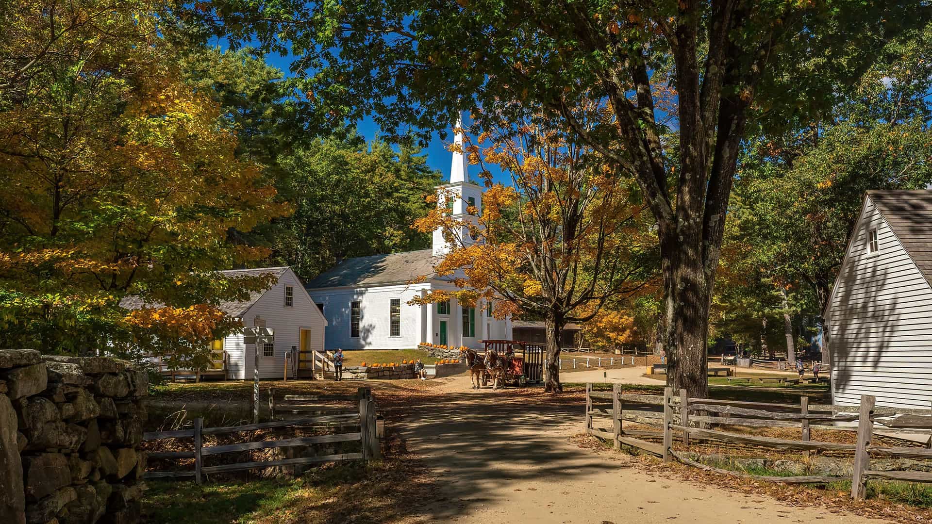"A scenic autumn view of the historic Old Sturbridge Village, featuring a white church with a steeple surrounded by colorful trees, in Massachusetts.  "