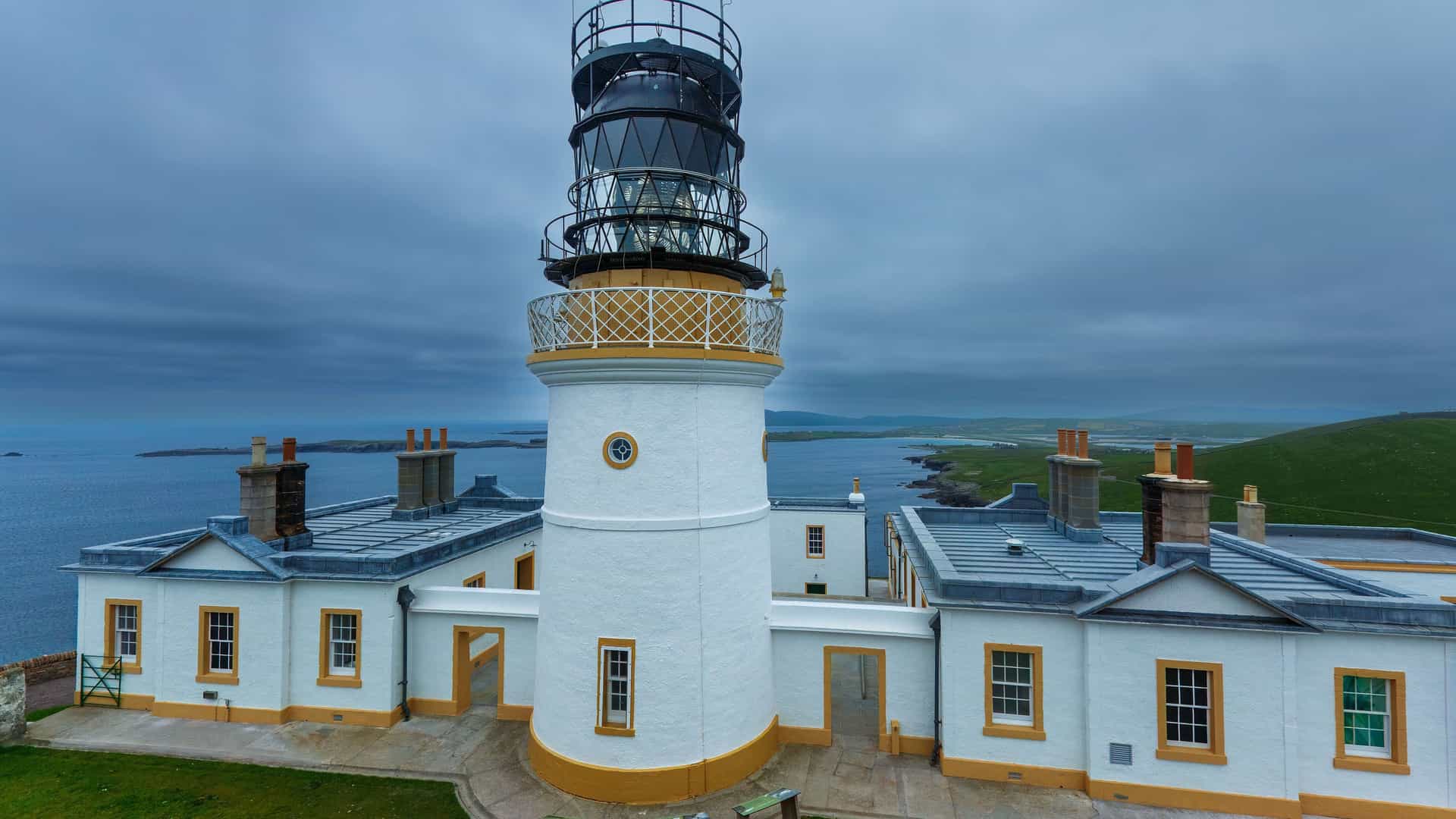 An aerial view of the Sumburgh Head Lighthouse and its surrounding buildings on a cliff overlooking the sea in Scotland. The sky is overcast with gray and white clouds.