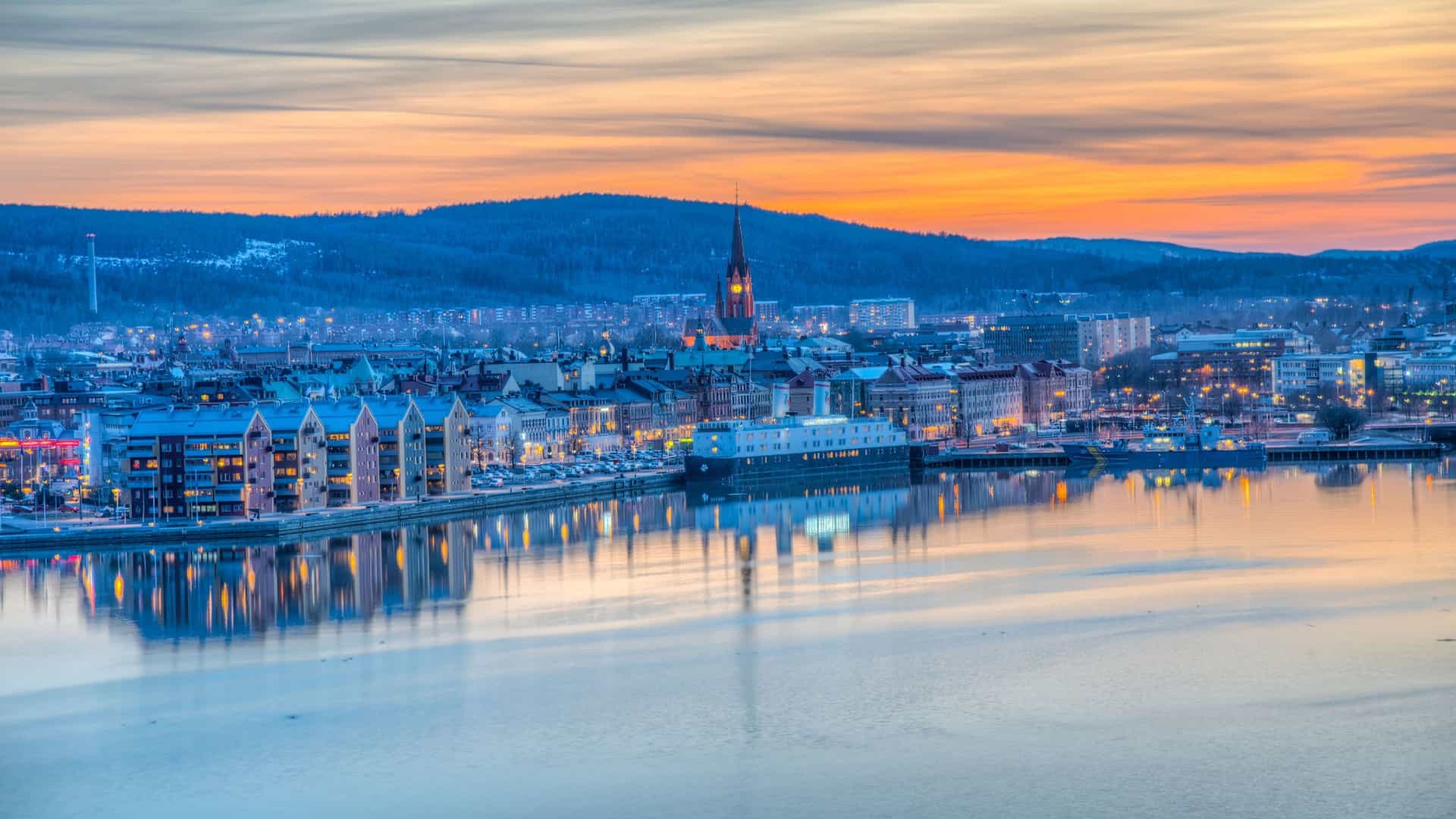 A scenic winter evening view of Sundsvall, Sweden, with the city lights reflected in the frozen water of the harbor and snow-covered hills in the distance.