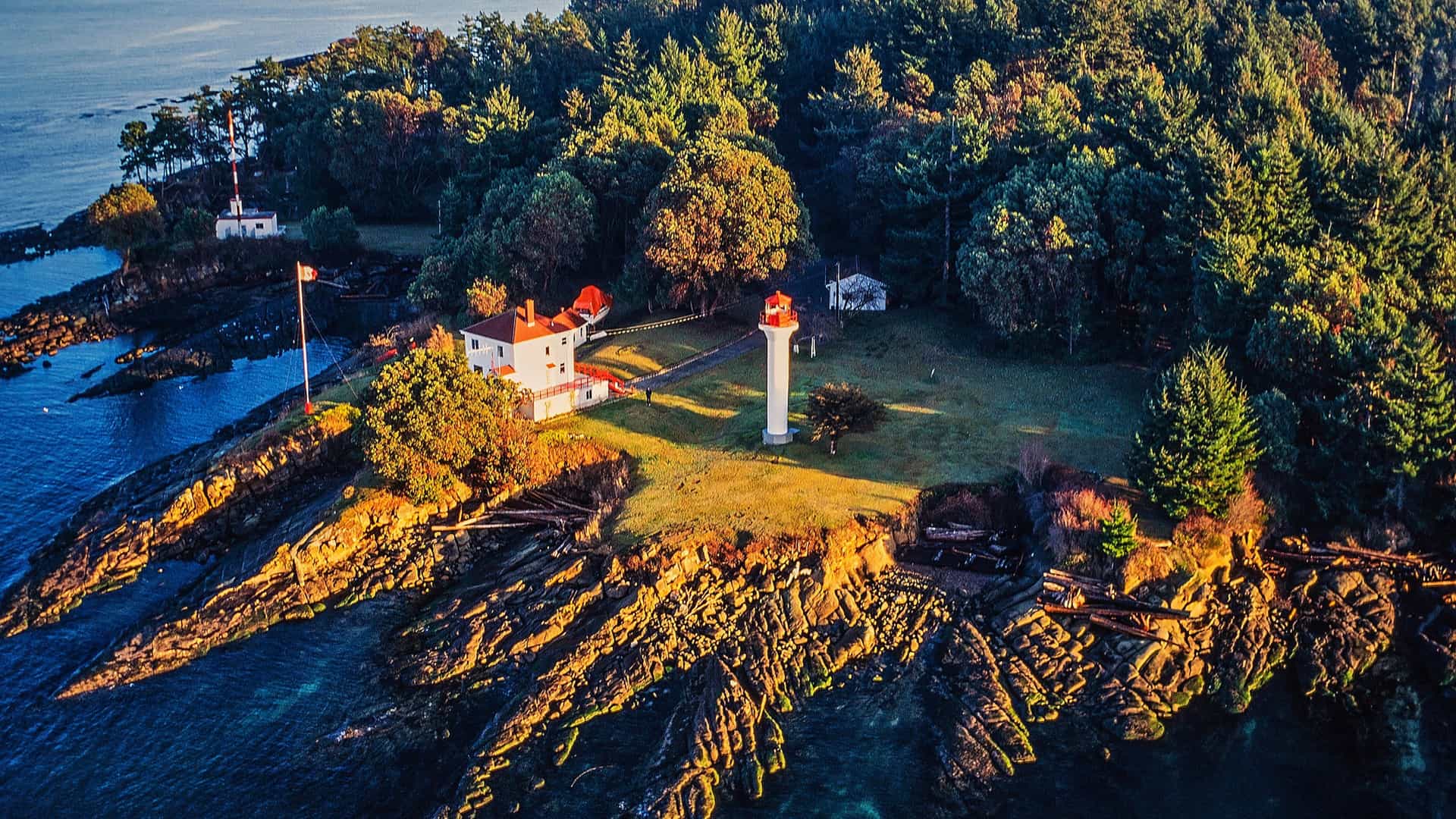 An aerial view of a white lighthouse and residence on a rocky, wooded peninsula jutting into the blue water of Swartz Bay.