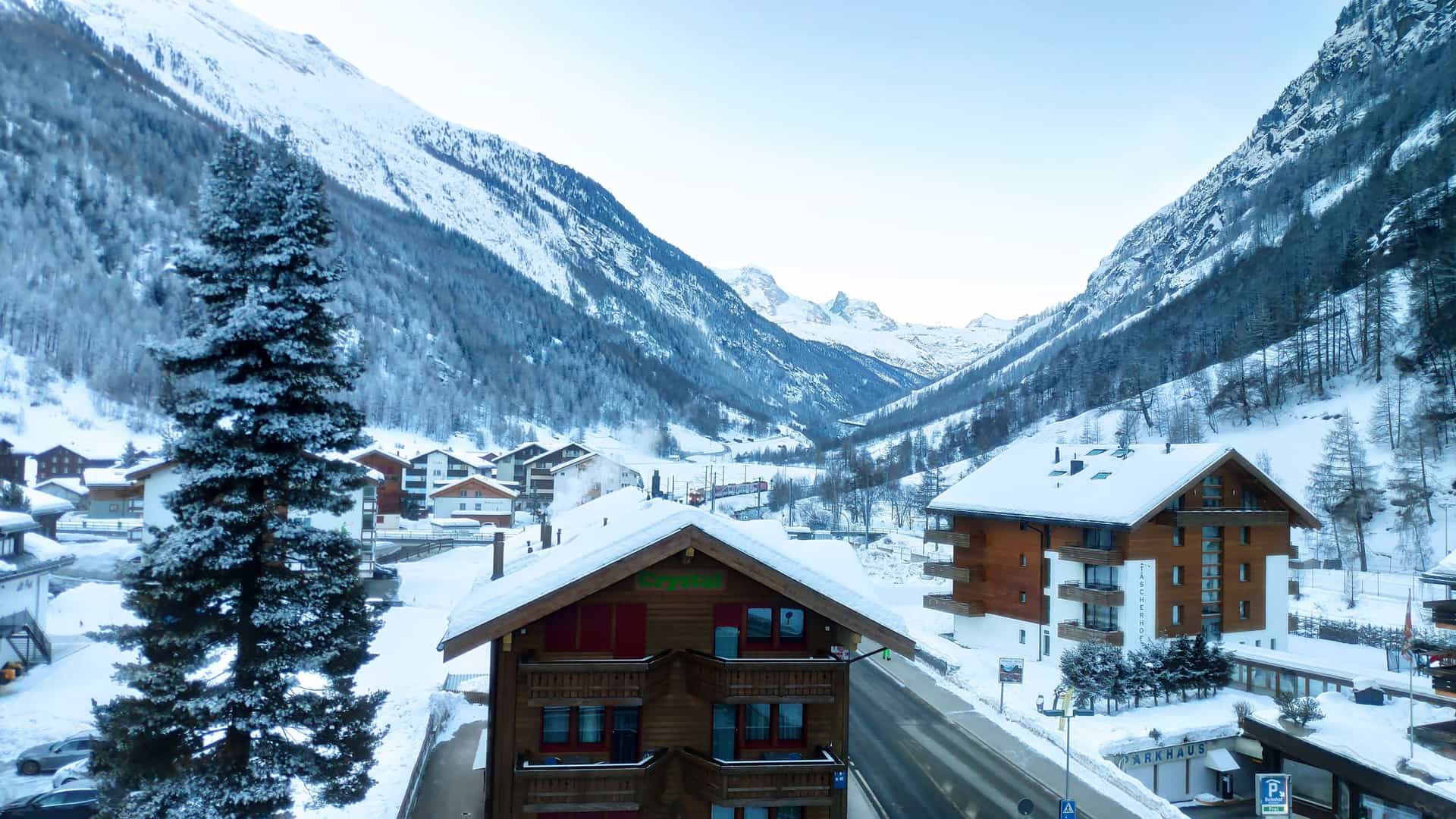 A picturesque winter view of the village of Täsch, Switzerland, with traditional wooden houses and snow-covered streets nestled in a valley surrounded by towering snow-capped mountains.