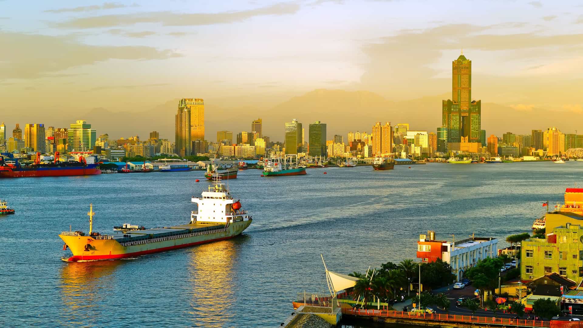 A wide-angle sunset view of the busy shipping port of Kaohsiung, Taiwan, with a large tanker and other ships sailing in the Taiwan Strait with the modern cityscape and high-rise buildings in the background.