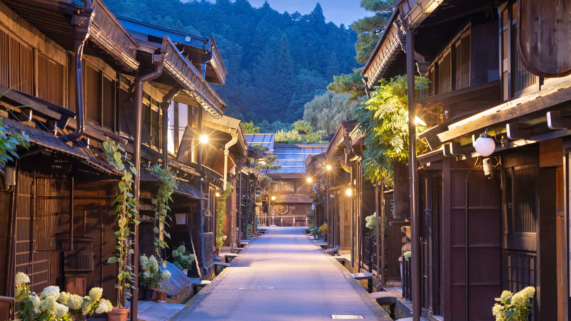 "A serene evening view of the historic district of Takayama, Japan, showing a narrow, empty street lined with traditional, dark wooden Japanese buildings and glowing lanterns.  "