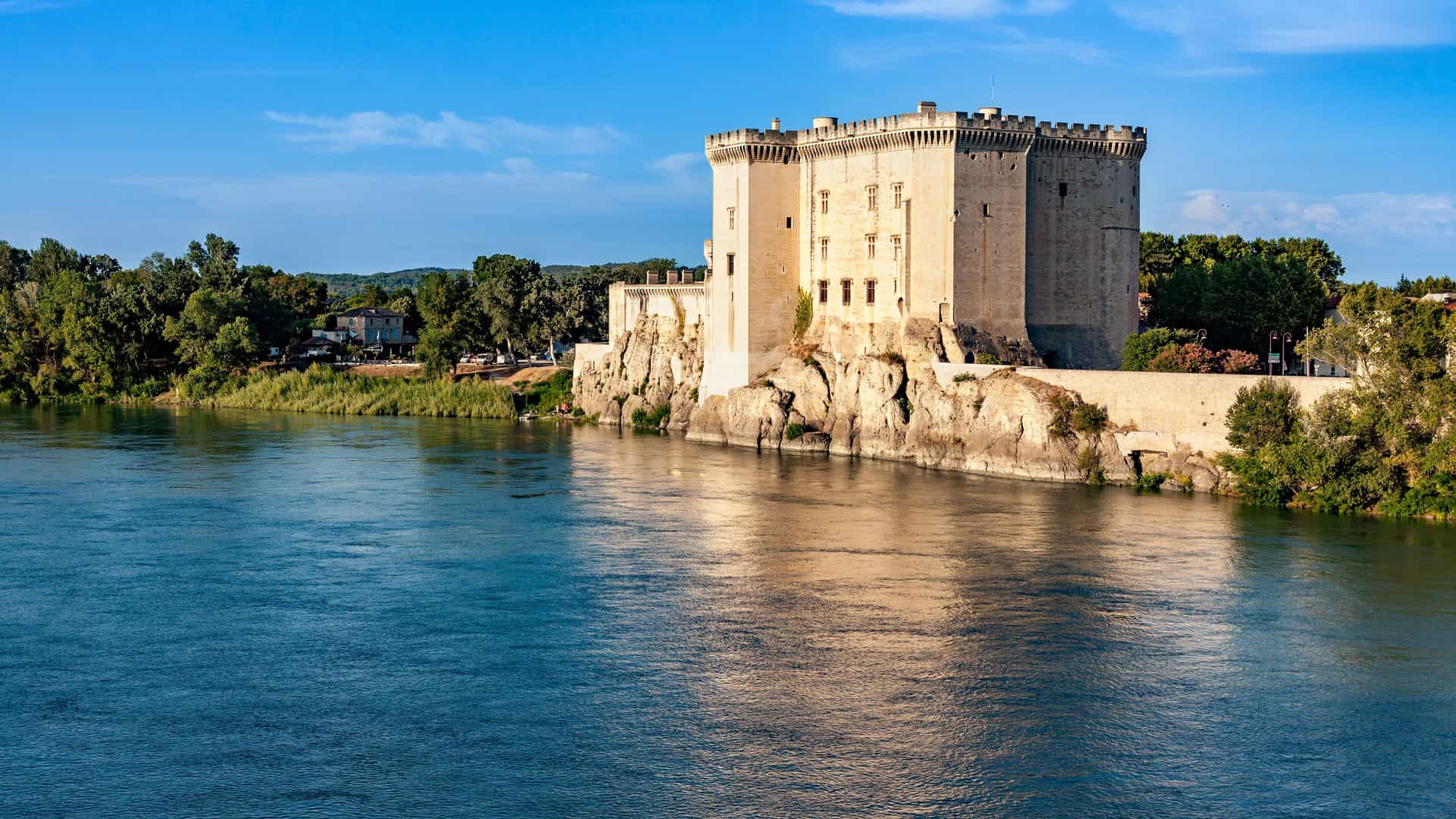 A stunning view of the medieval Château de Tarascon, a large stone fortress with towers and ramparts, rising from the banks of the Rhône River in Tarascon, France.