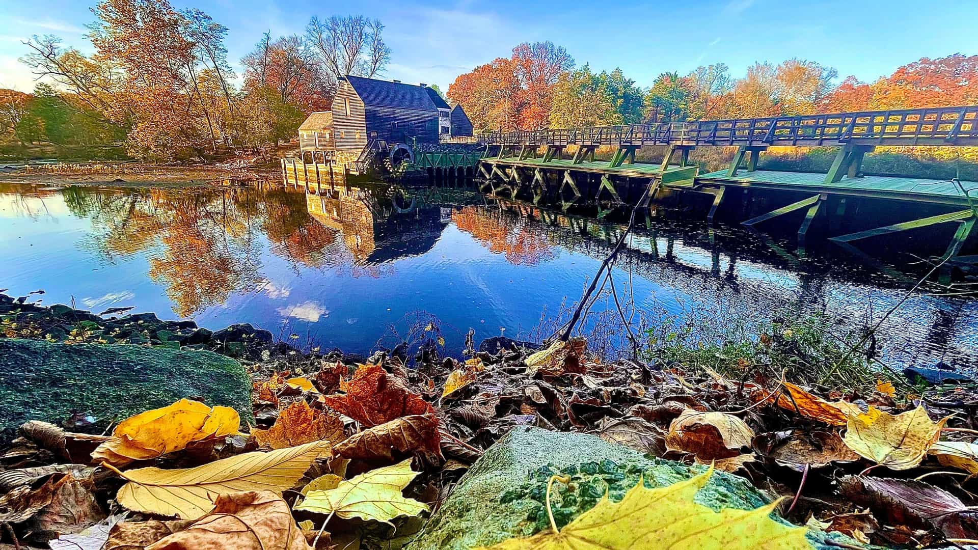 "A vibrant autumn scene of the historic Philipsburg Manor, an 18th-century grist mill with a water wheel and a wooden bridge, reflected in the still water in Tarrytown, New York.  "