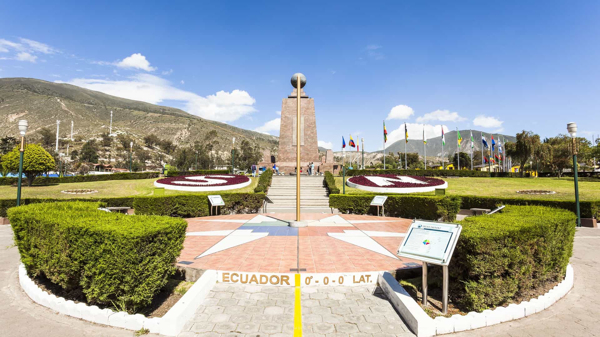 A view of the Mitad del Mundo monument at the Equator in Ecuador, with a circular plaza, garden beds, and a tall stone monument with a globe on top.