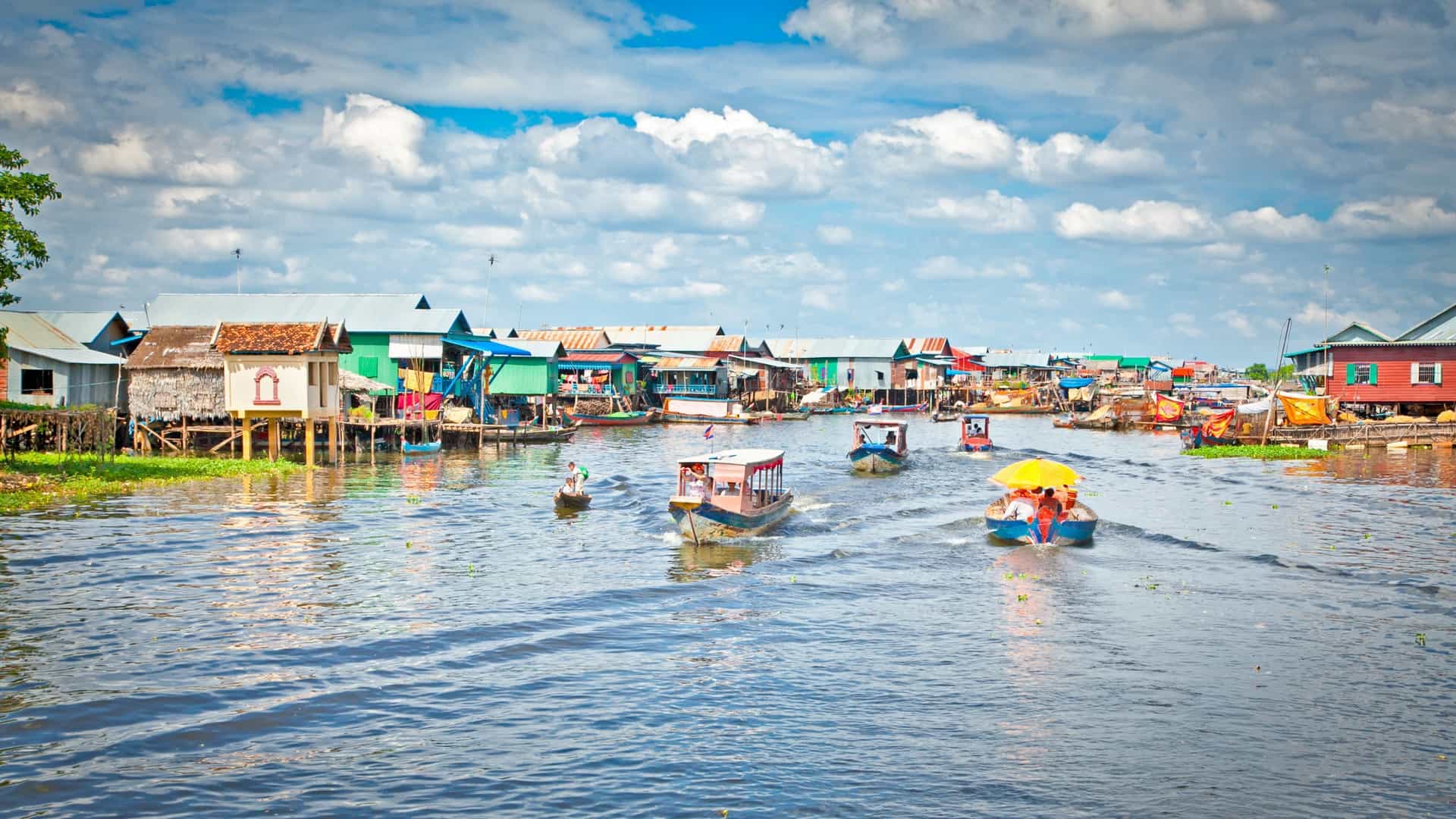A view of the Komprongpok floating village on Tonle Sap Lake, Cambodia. The community consists of numerous wooden houses and structures built on floating pontoons or barges, with small boats navigating the calm water between the dwellings.