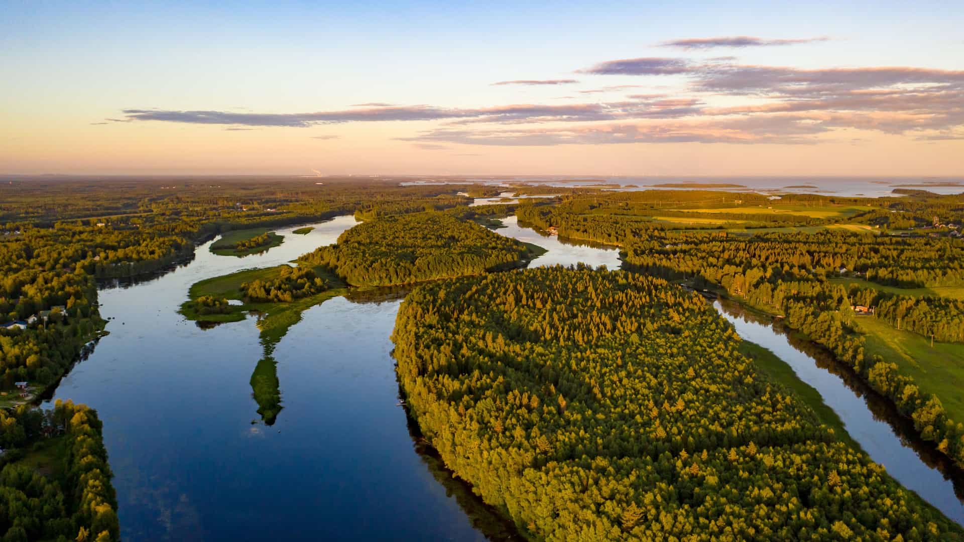 "An aerial view of the Tornio River winding through a lush green landscape of forests and fields in Finland, with the calm water reflecting the beautiful sunset sky.  "