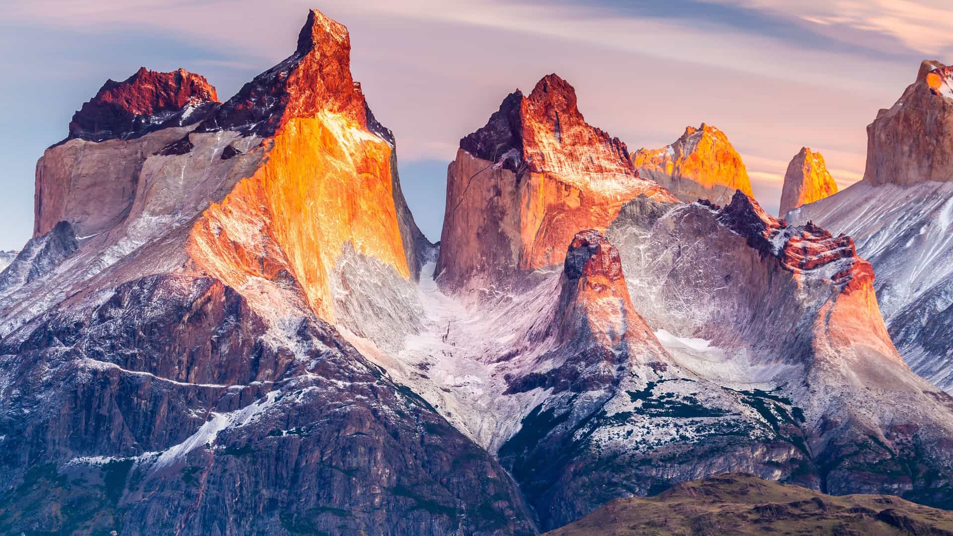 "A striking view of the towering granite peaks of the Torres del Paine Massif in Chile, with the sun's warm light illuminating the jagged rock formations and a dusting of snow.  "