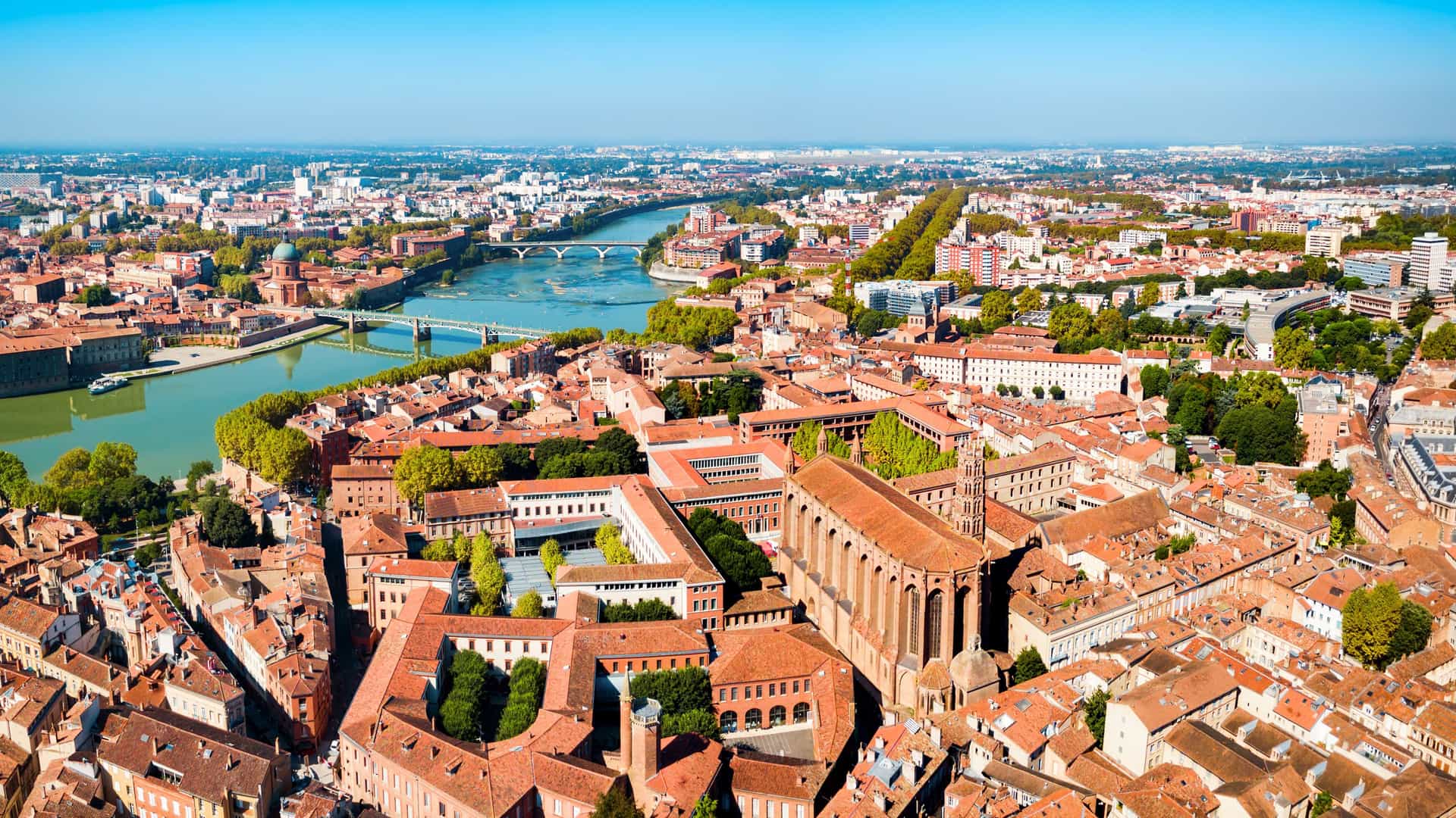 "An aerial panoramic shot of Toulouse, France, showing the city's signature red-brick buildings along the Garonne River and its bridges, with the beautiful Basilique Saint-Sernin church prominently featured.  "