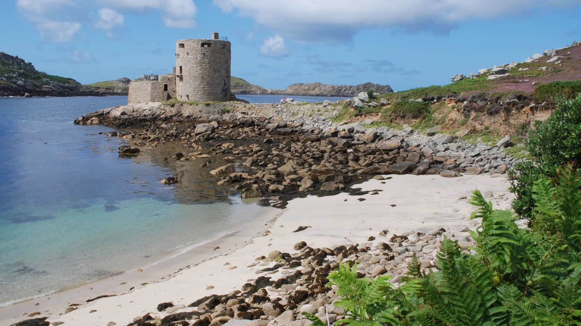A scenic view of Cromwell's Castle, a historic stone circular fortress ruin, on the rocky coastline of Tresco, with clear turquoise water and a small sandy beach.