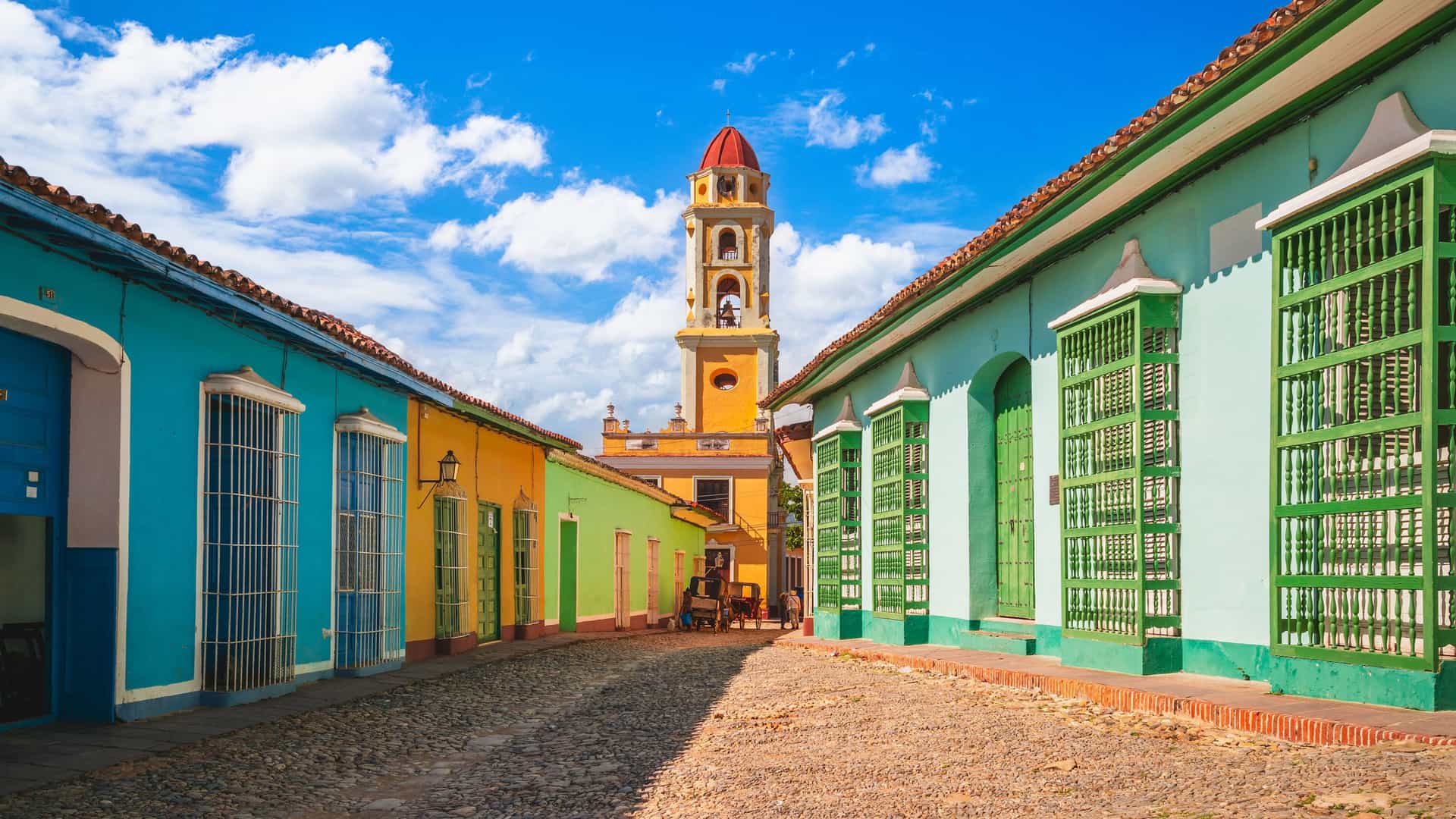 "A cobblestone street in Trinidad, Cuba, lined with colorful historic buildings in shades of blue, green, and yellow, leading towards the iconic bell tower of the Iglesia y Convento de San Francisco.  "