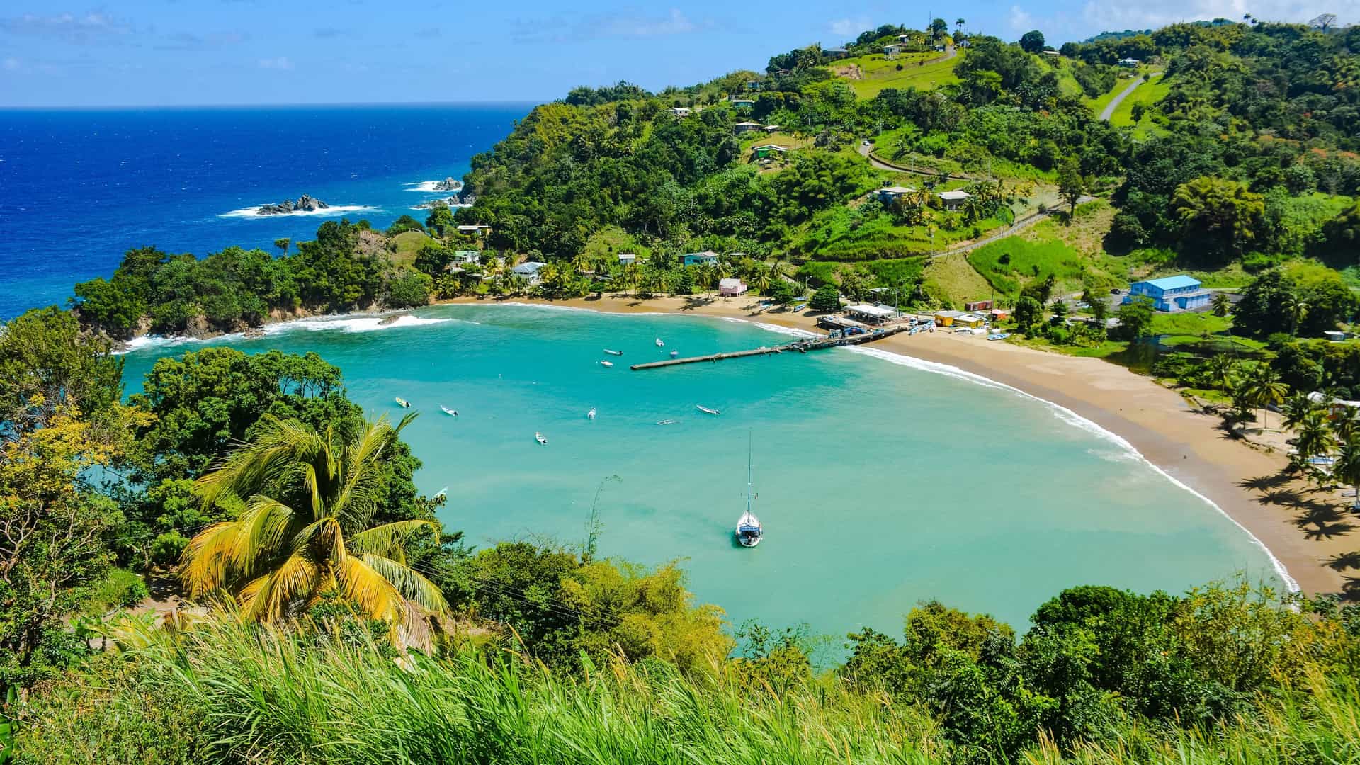 A scenic aerial view of a pristine beach cove in Trinidad, with turquoise water, a fishing pier, and lush green hillsides dotted with houses.