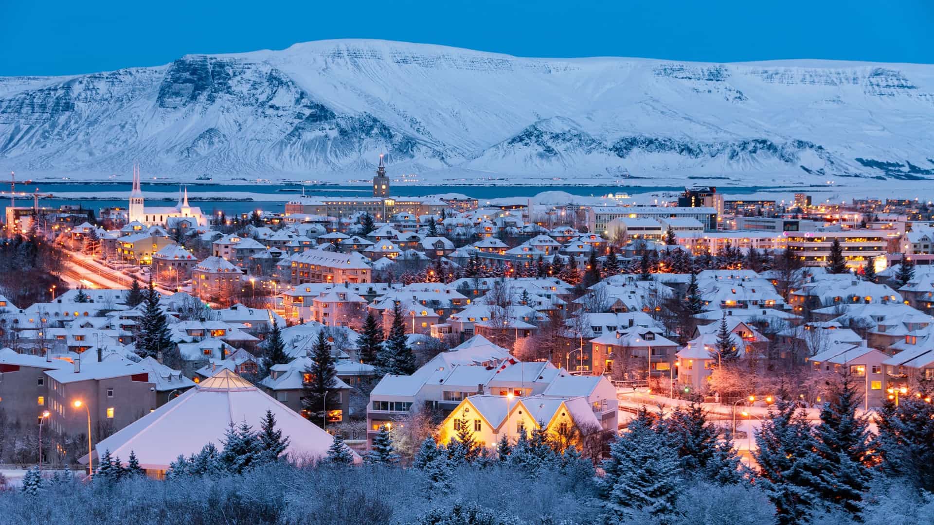 A snowy evening view of Reykjavik, Iceland, with illuminated buildings and the Hallgrímskirkja church spire visible, set against the backdrop of snow-covered Mount Esja and the water.