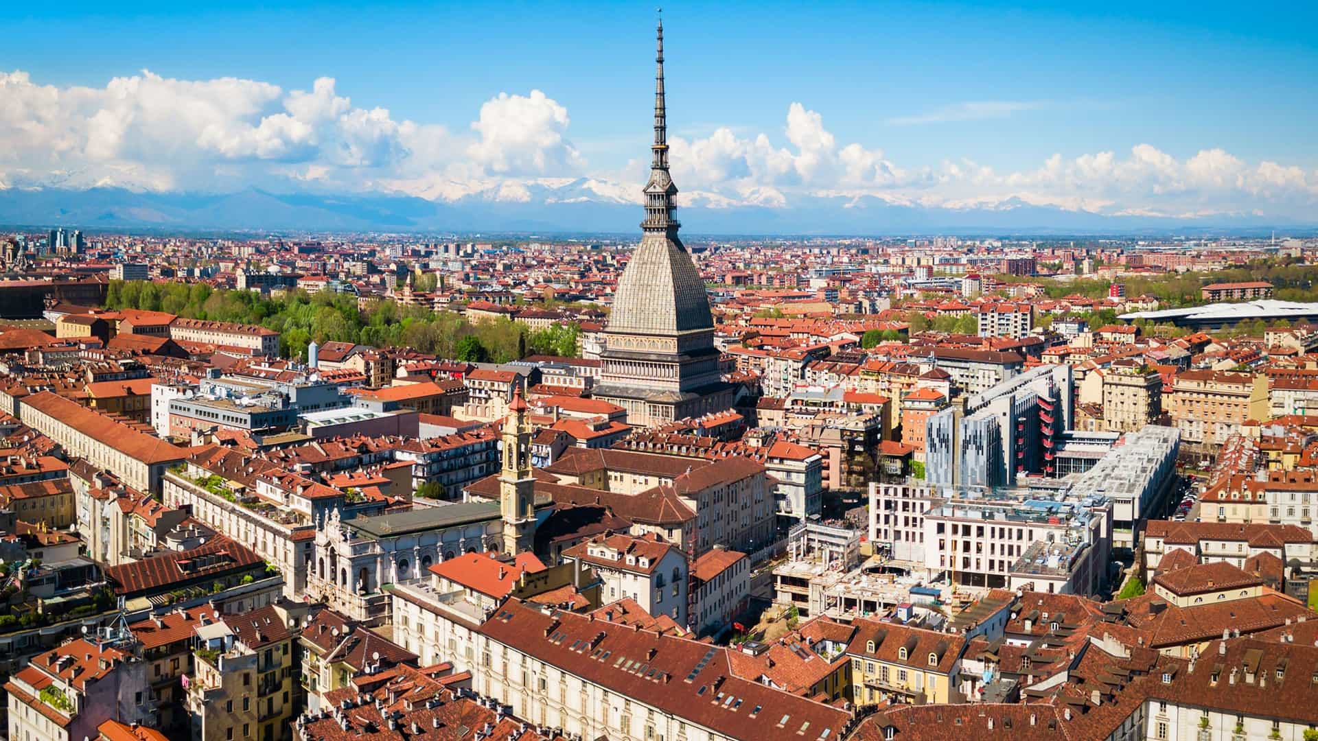 "An aerial panoramic shot of Turin, Italy, highlighting the iconic Mole Antonelliana spire rising above the cityscape of red-tiled roofs, with the snow-capped Alps in the background.  "