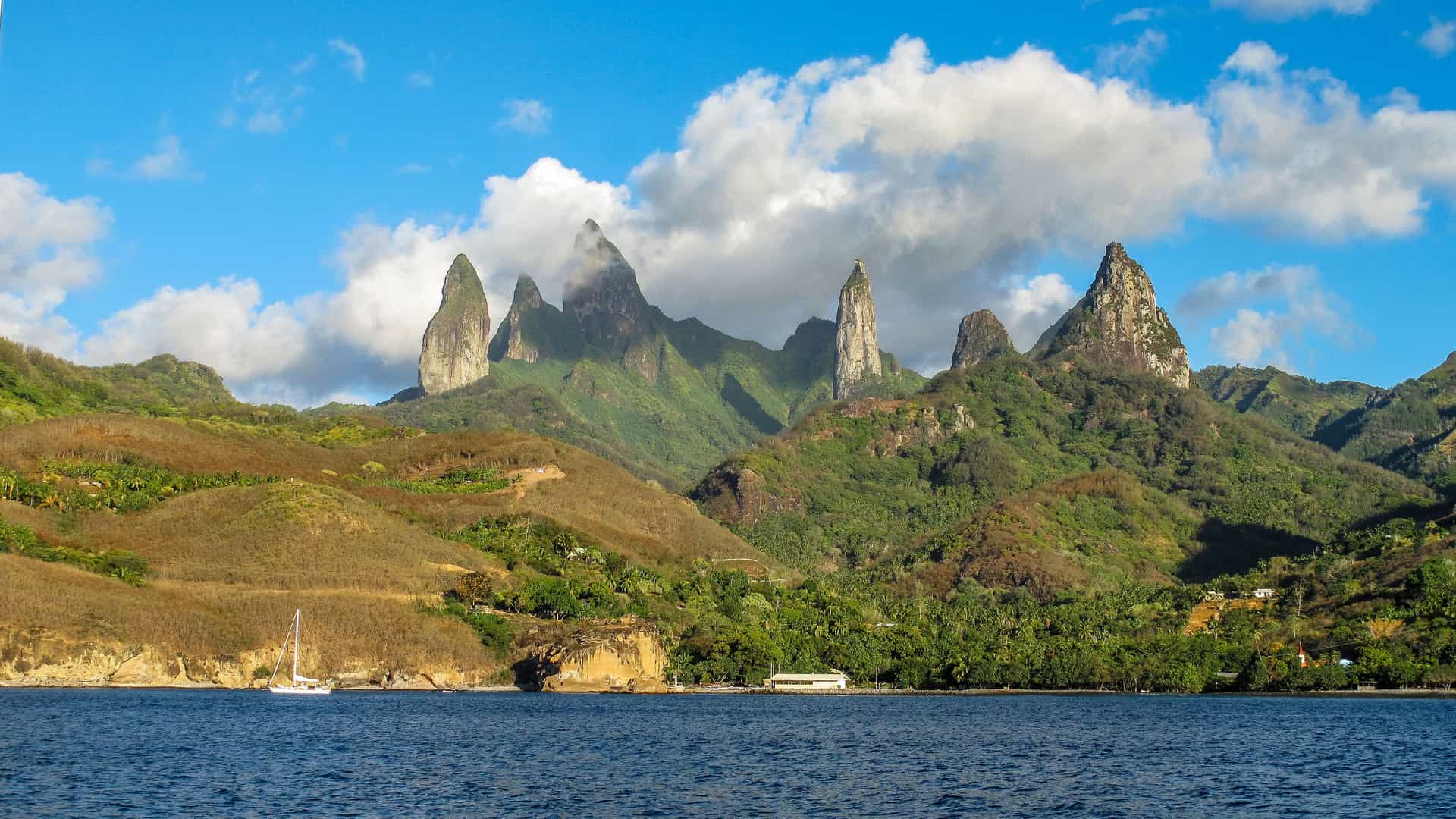 A dramatic view of the sharp, volcanic basaltic spires of Ua Pou, a landmark in the Marquesas Islands, rising from the lush green hills above a tranquil blue bay.