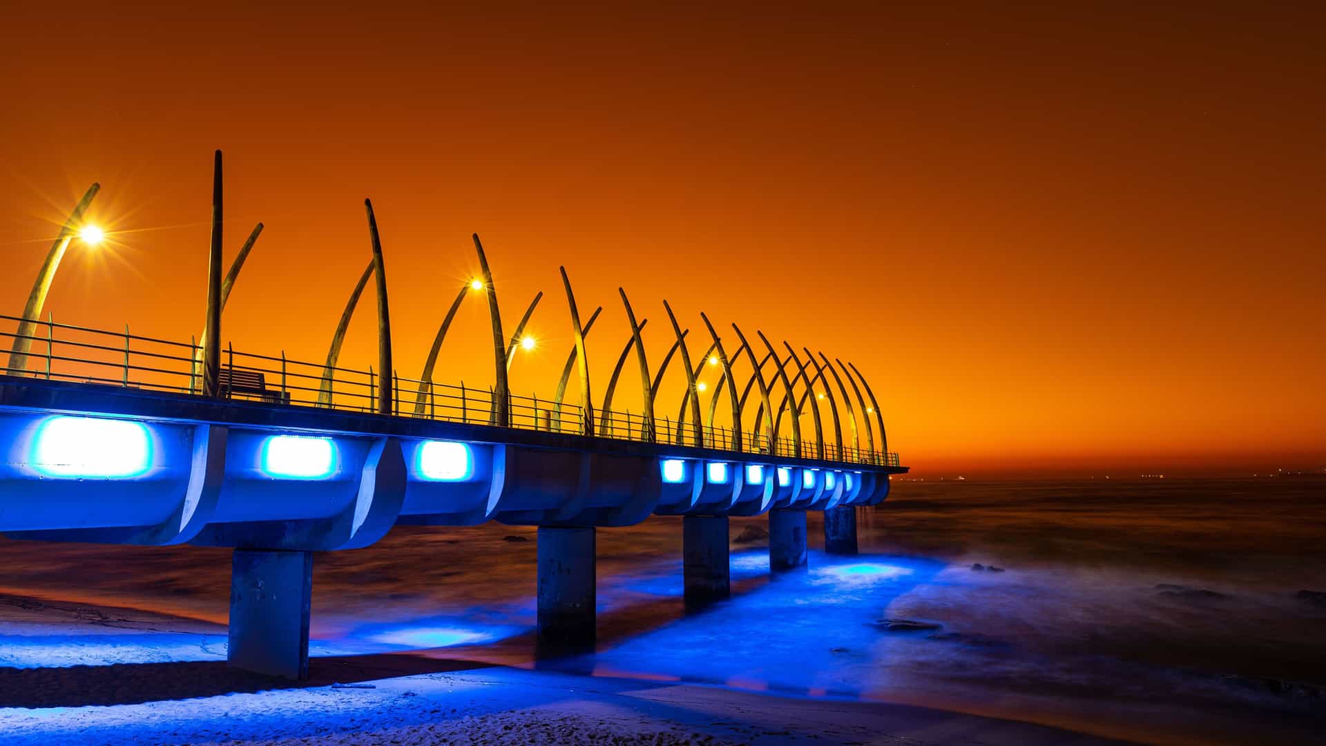 "A striking nighttime view of the Whalebone Pier in Umhlanga, South Africa, illuminated with bright blue lights, with the orange glow of the rising sun or the city lights reflecting off the night sky.  "