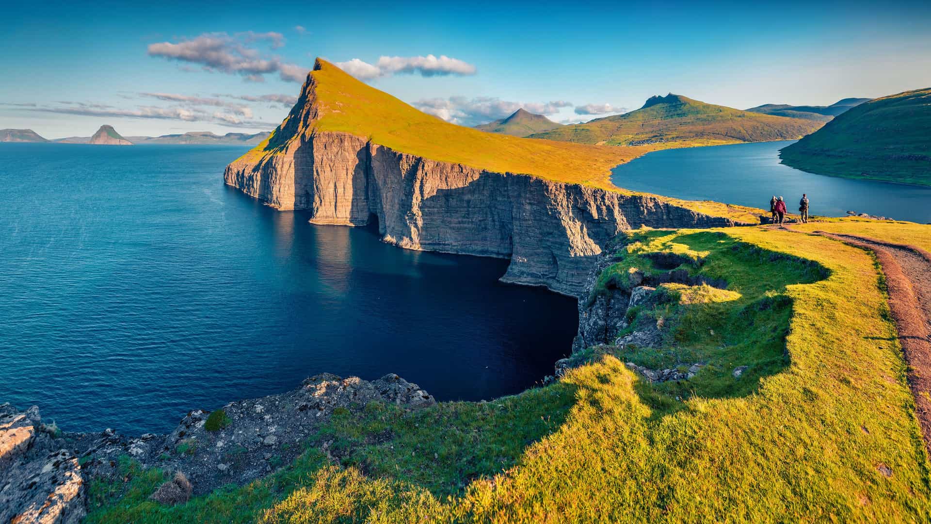 "A breathtaking panoramic shot of Lake Sørvágsvatn, also known as ""The Lake Above the Ocean,"" on the island of Vágar in the Faroe Islands, Denmark. The image shows the steep, grassy cliffs and calm water with a hiking trail and a person in the foreground.  "