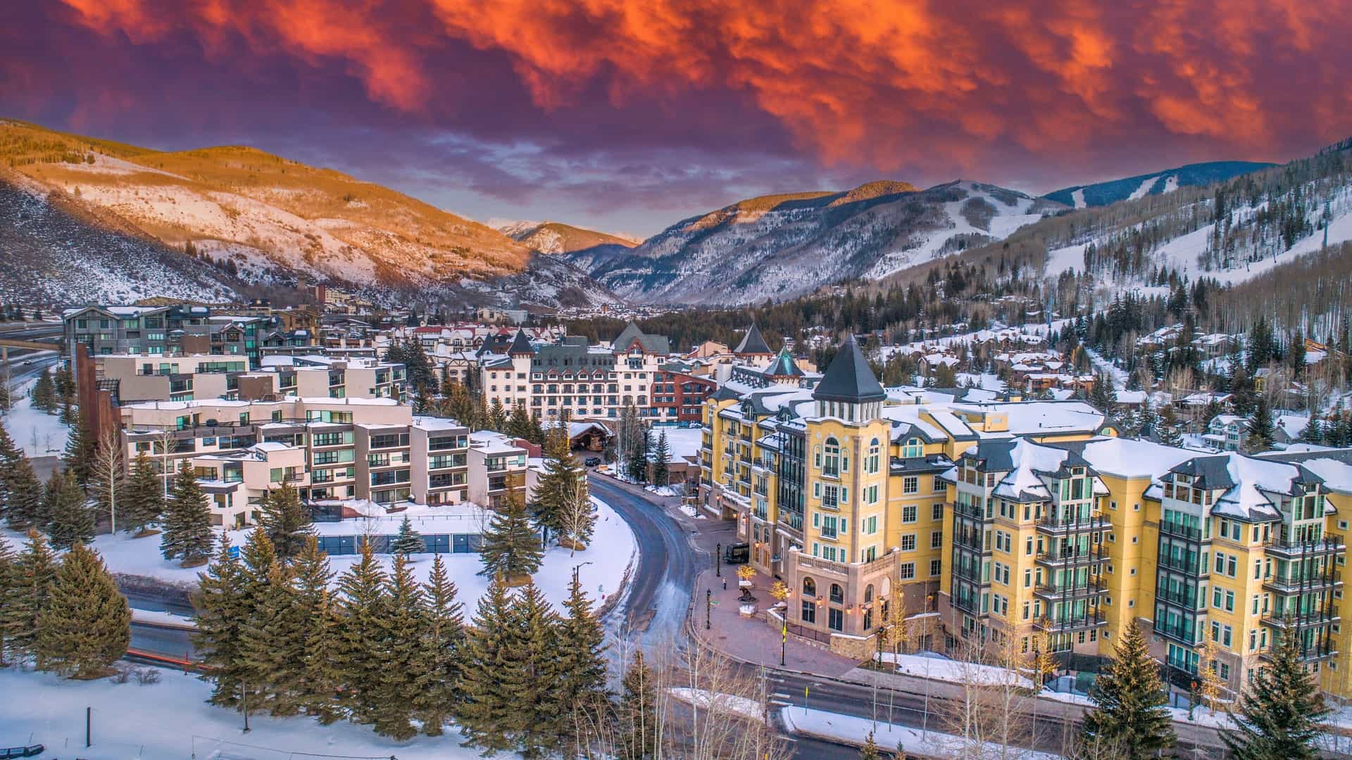 An alpine village with European-style architecture is nestled in snow-covered mountains under a dramatic orange sunset in Vail, Colorado.