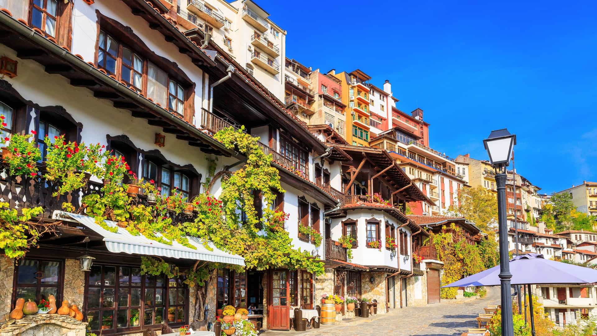 A picturesque street scene in Veliko Tarnovo, Bulgaria, showcasing traditional Bulgarian revival houses with stone and wood construction, balconies, and hanging flowers under a clear blue sky.
