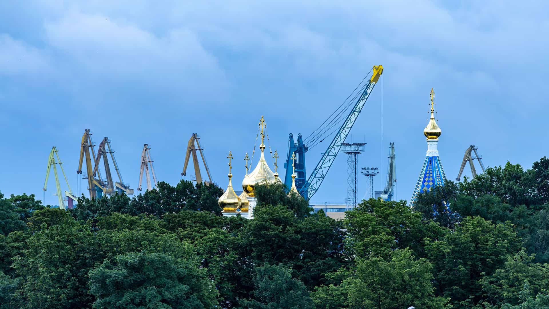 A striking contrast between old and new in Ventspils, Latvia, with the golden domes of an Orthodox church towering above green trees, set against the backdrop of industrial cranes at the city's port.