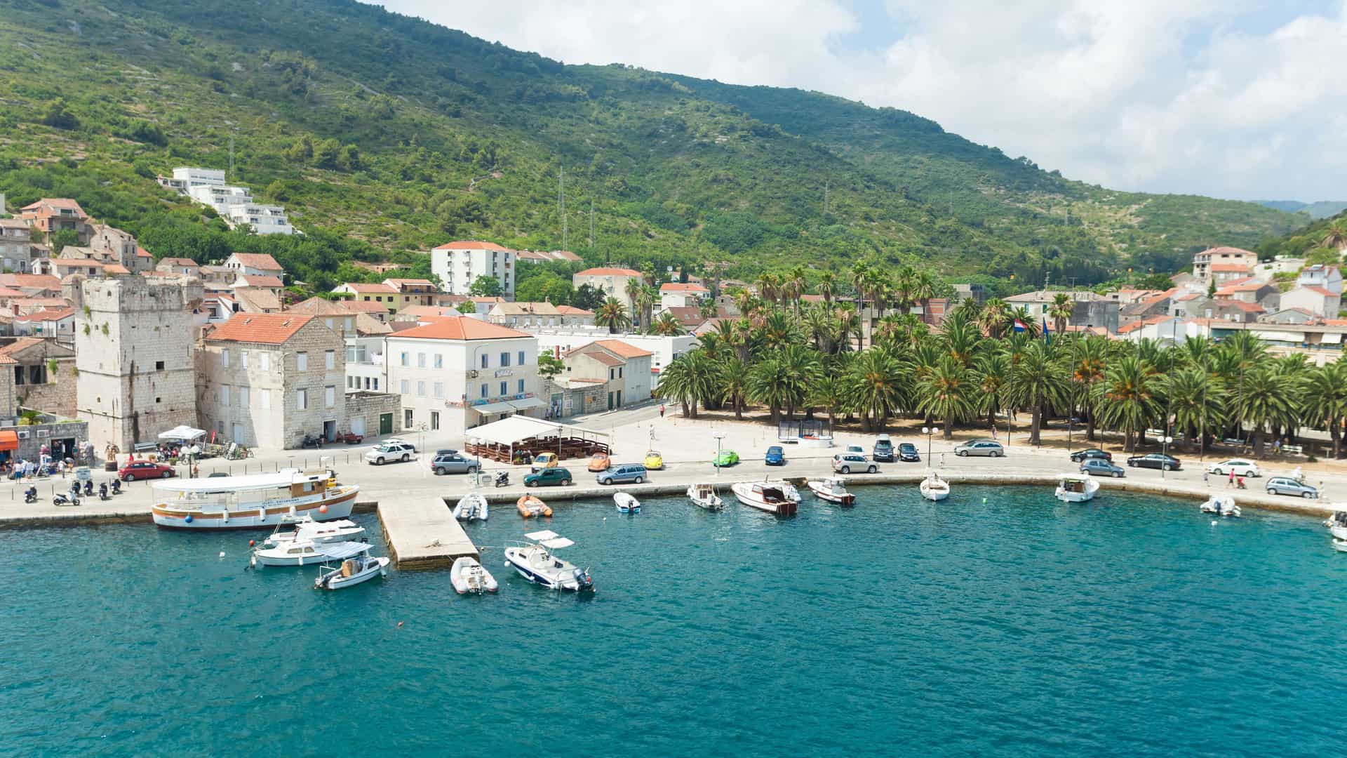 A scenic view of Vis harbor in Croatia, with small boats docked at the pier, surrounded by historic buildings and a lush green hillside.