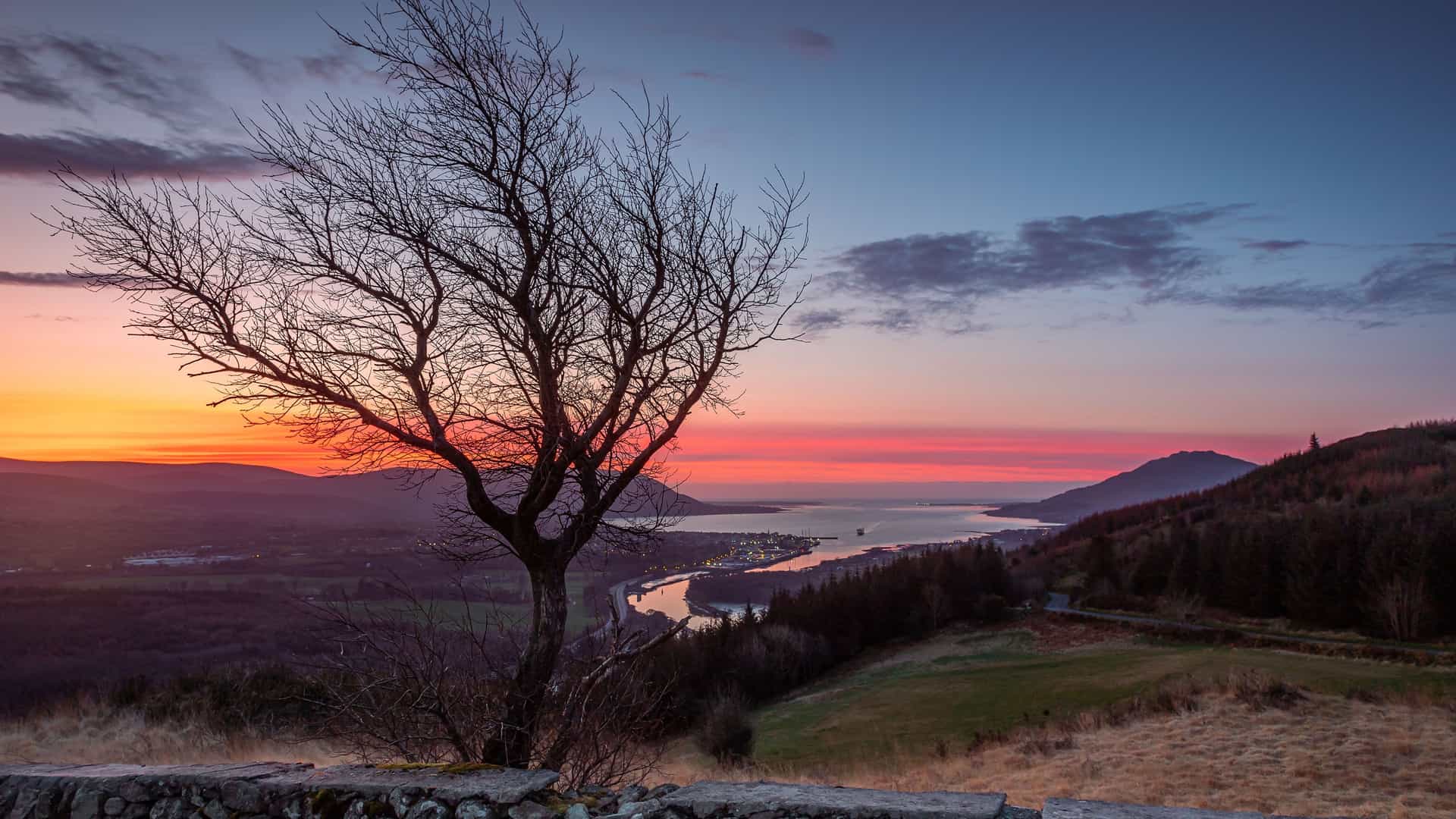 A scenic elevated view of the town of Warrenpoint in Northern Ireland, nestled on the shores of Carlingford Lough and surrounded by Mourne Mountains during a vibrant sunset.