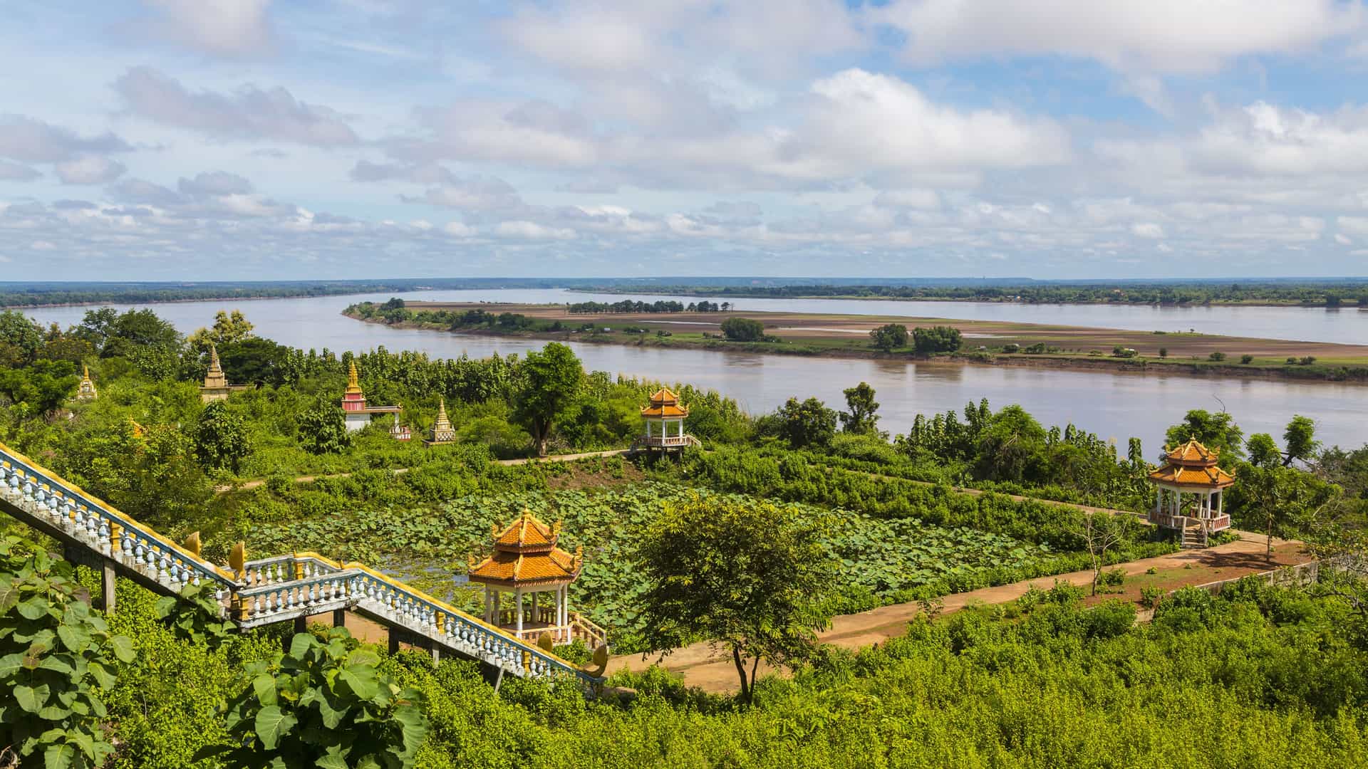 A scenic vista including Wat Hanchey, a hilltop temple overlooking the vast Mekong River in Cambodia, surrounded by vibrant green foliage and small religious structures. 