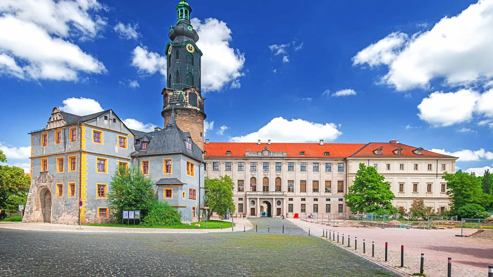 "A sunny, wide-angle shot of the historic Weimar City Palace in Germany, showcasing its large clock tower and Renaissance-style architecture against a vibrant blue sky.  "