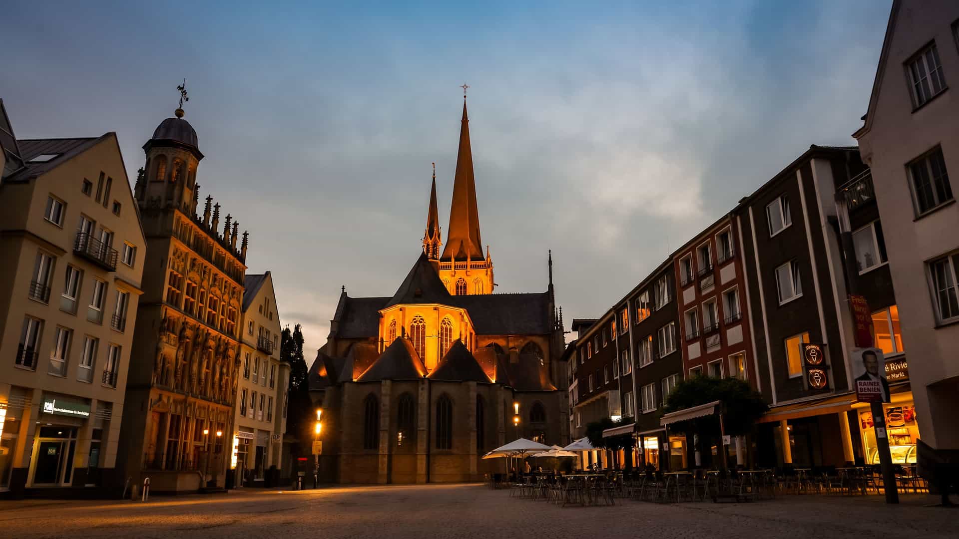 A view of a large church at night with its spires lit up in the center of the town square in Wesel, Germany. The surrounding buildings are also illuminated, and there are empty tables and chairs from outdoor restaurants.