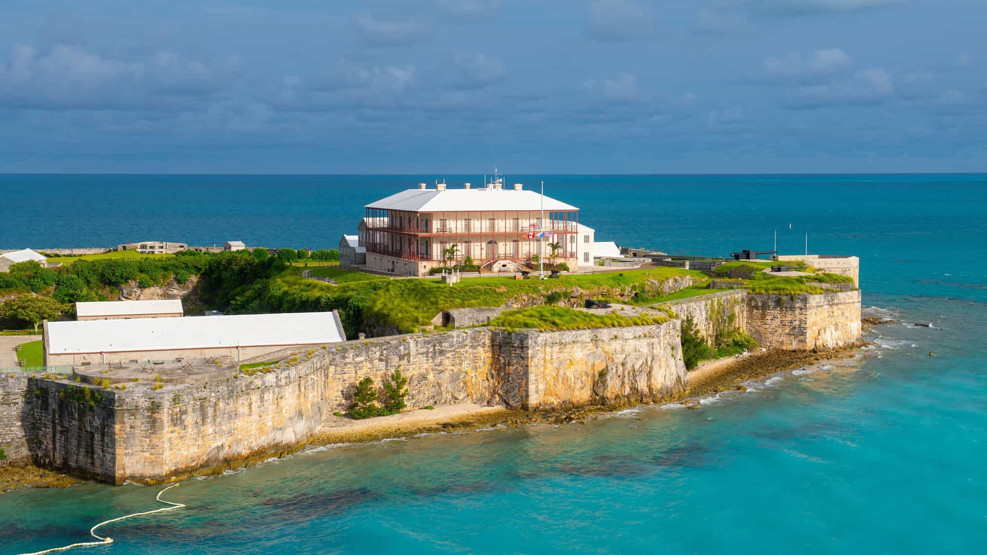 A view of a historic, two-story building with a white roof on a fort at the Royal Naval Dockyard, with the clear blue ocean surrounding the West End of Bermuda.